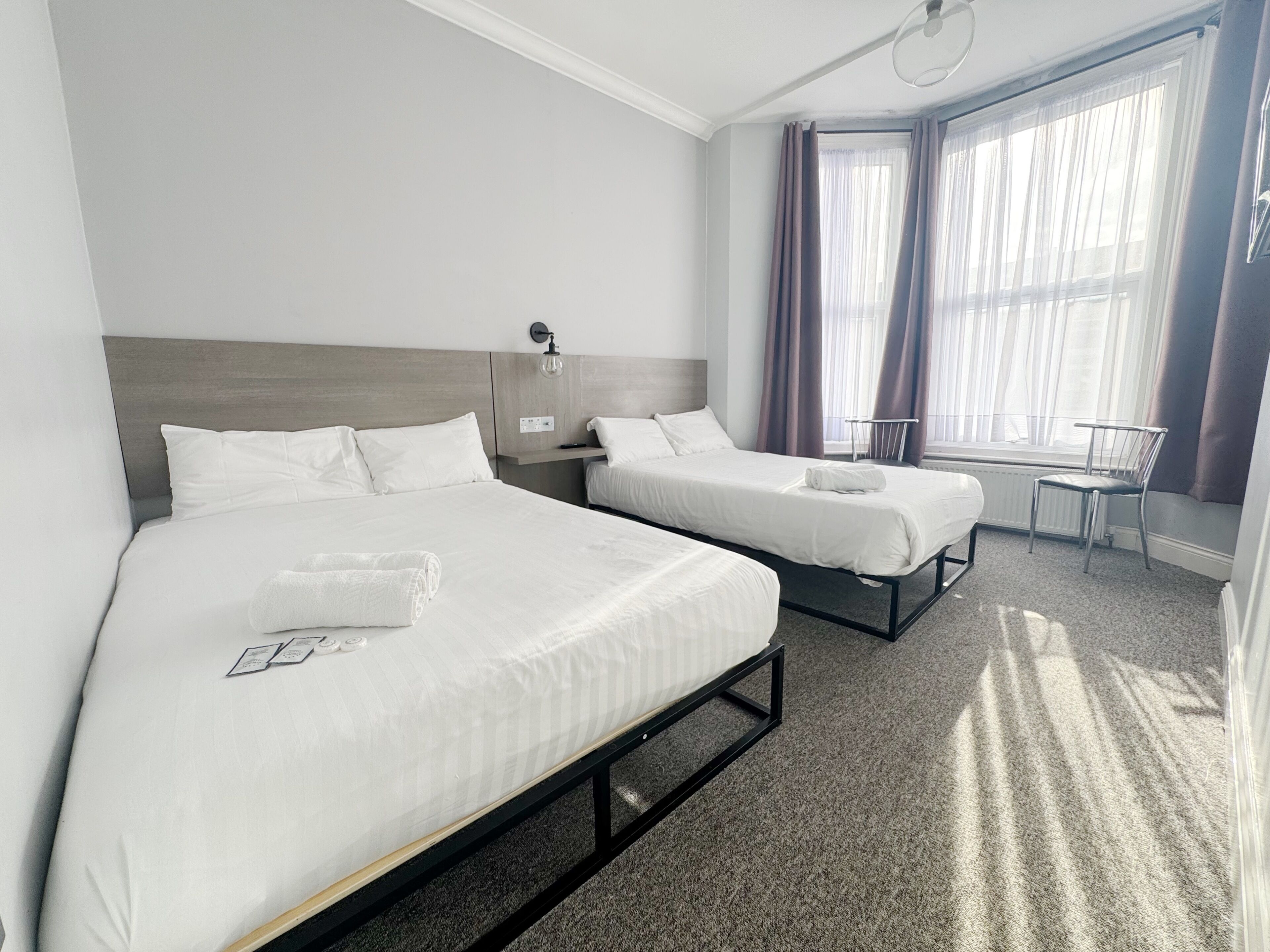 A hotel room with two beds, a window with curtains, a chair, and a radiator.