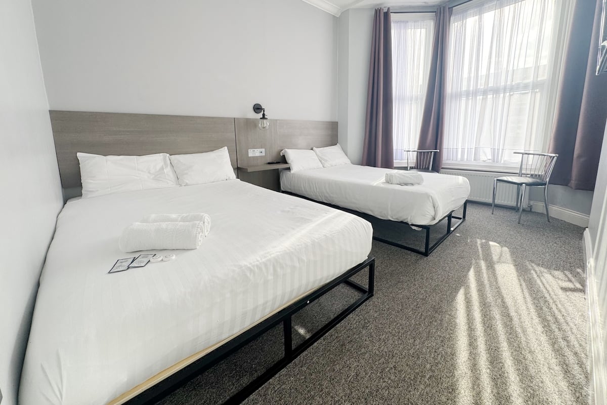 A hotel room with two beds, a window with curtains, a chair, and a radiator.