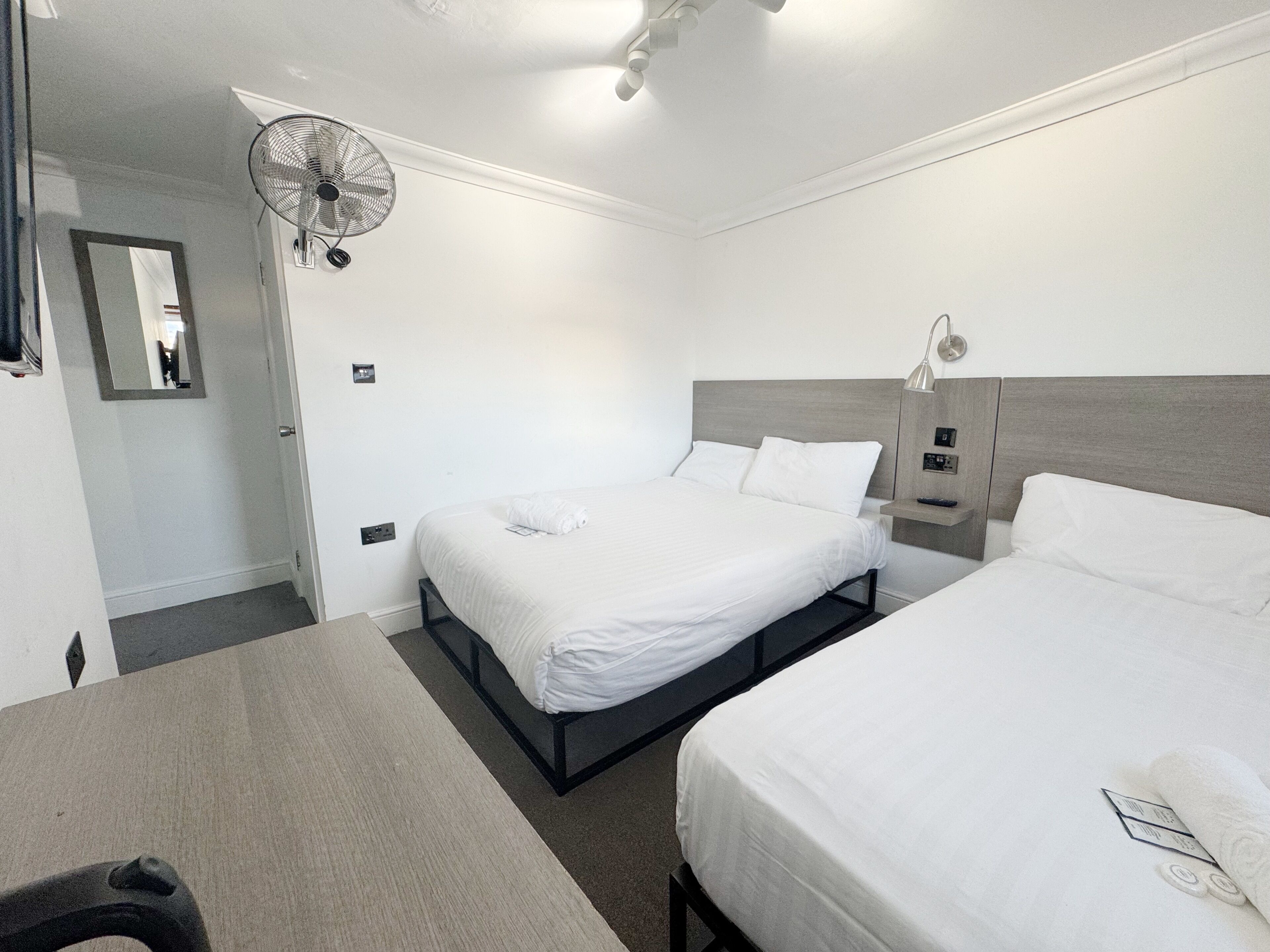 Ett hotellrum med två sängar, ett skrivbord, en spegel, en takfläkt och ett nattduksbord.