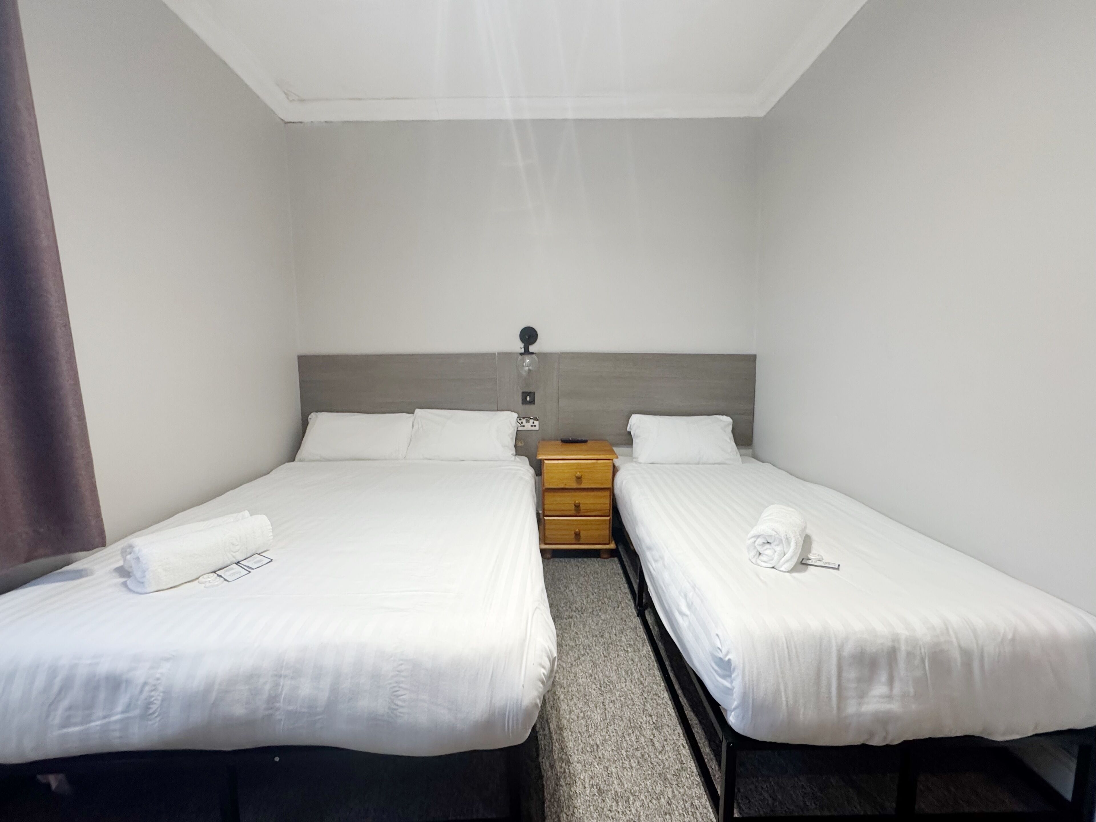 A hotel room with two single beds, a wooden nightstand, a wall socket, and a ceiling light fixture.