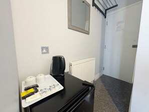 A hotel room with a black desk, a coffee maker, a tray with condiments, a mirror, a radiator, and a door with a sign. - Plaza Hotel Hammersmith (London)