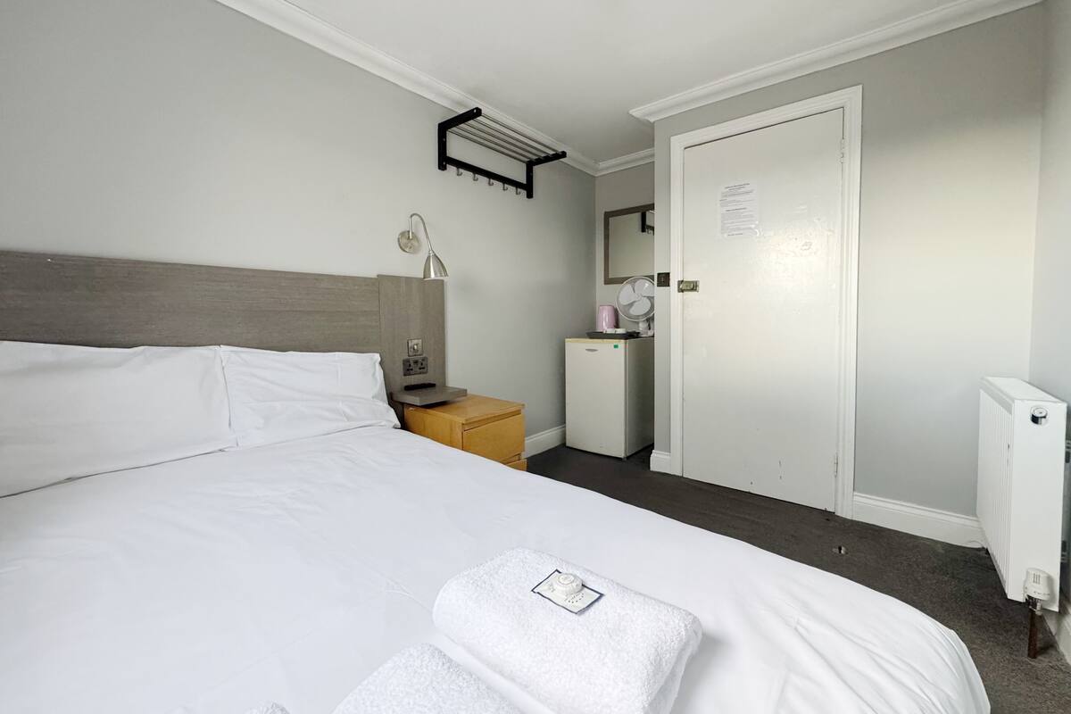 A hotel room with a bed, a bedside table with a lamp, a small refrigerator, a mirror, and a door.