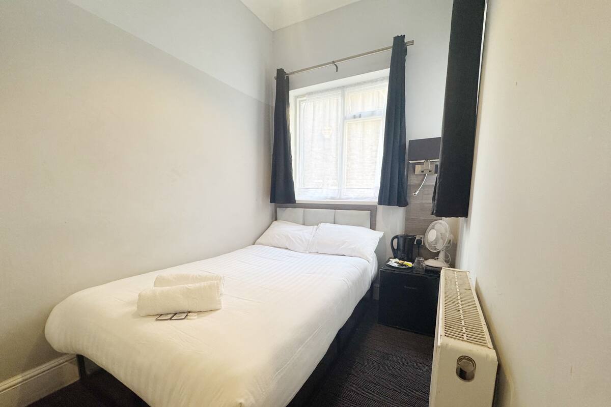 A hotel room with a bed, a bedside table with a fan, a heater, and a window with curtains.