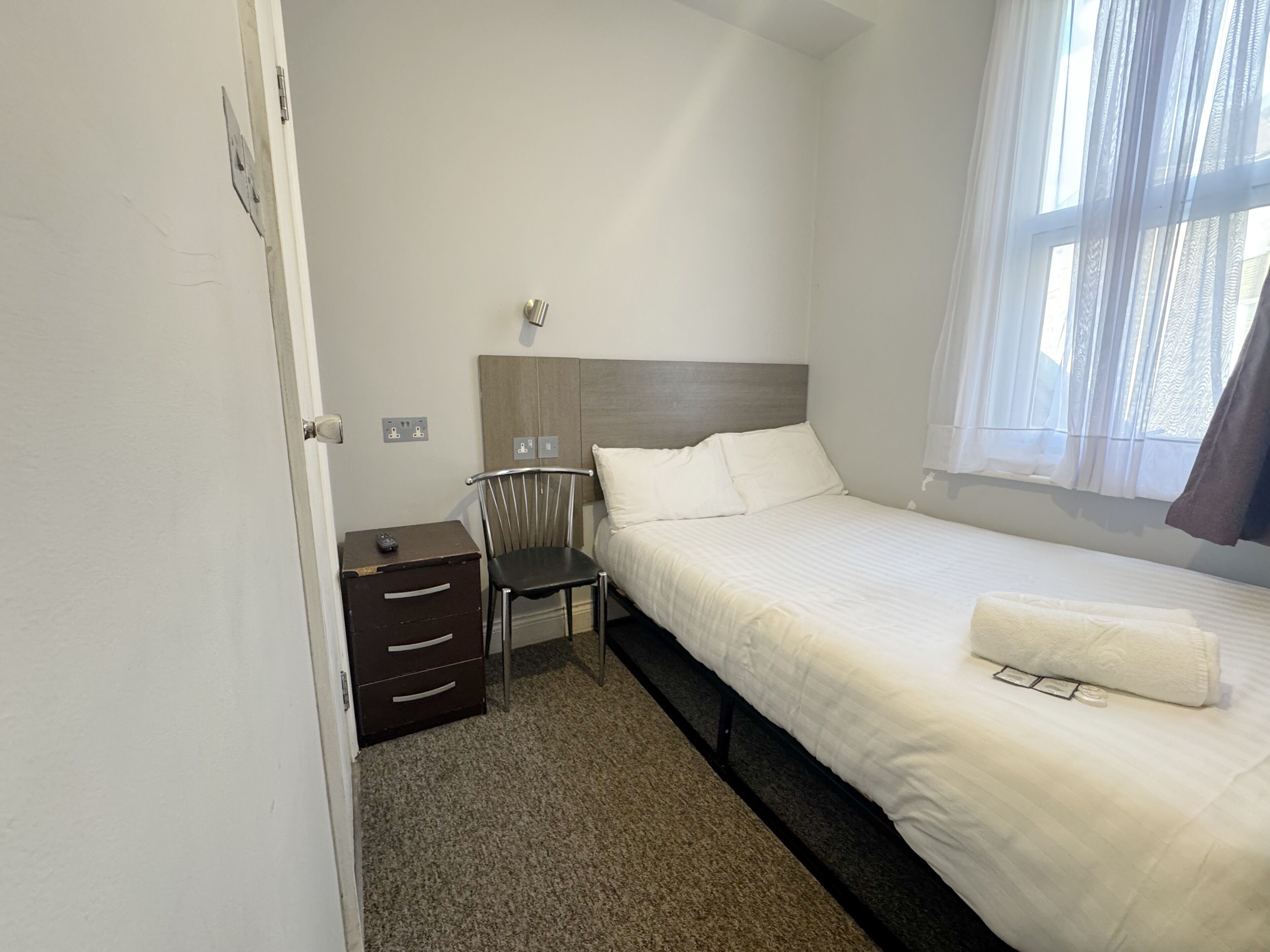 A hotel room with a bed, a chair, a nightstand, and a window with curtains.