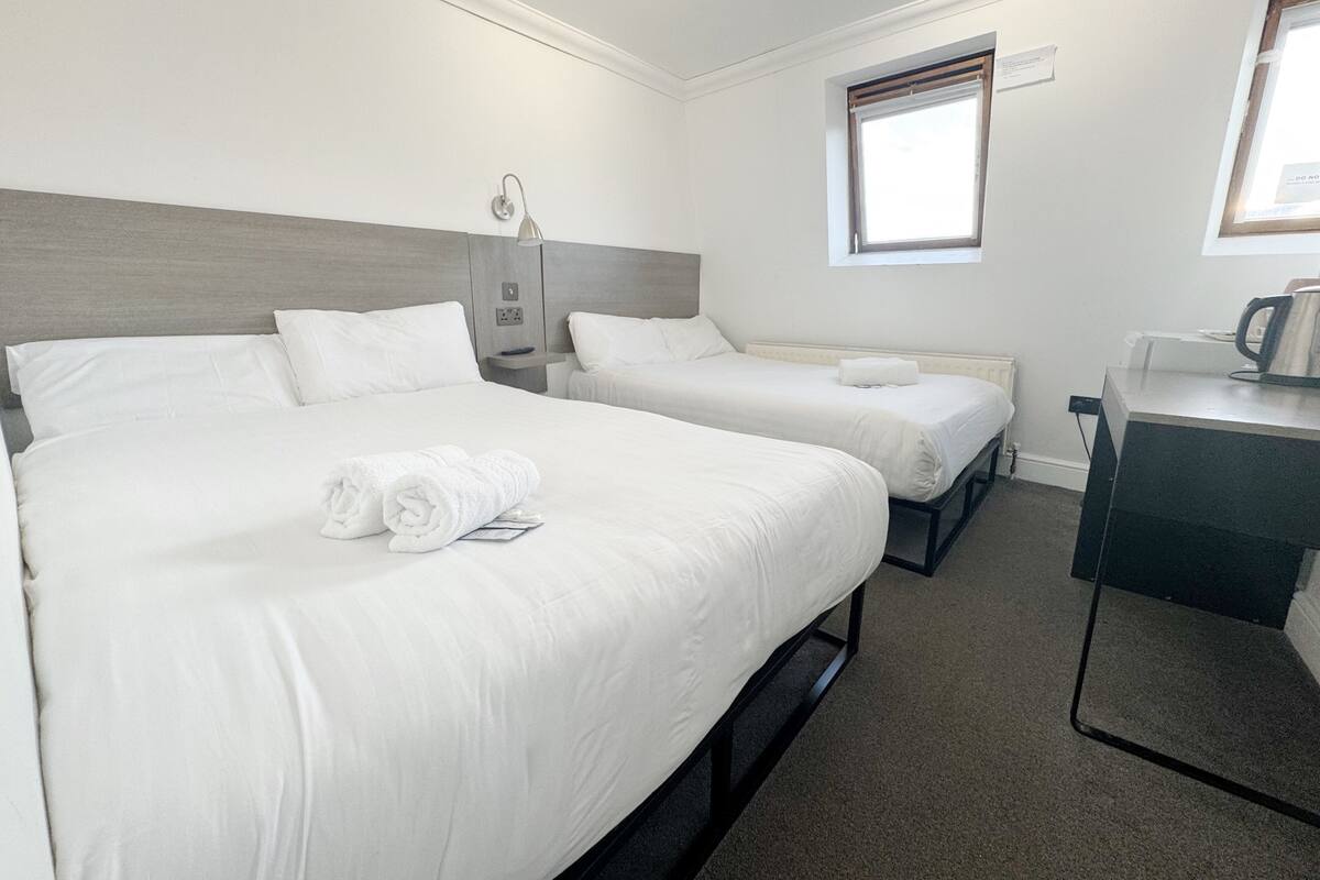 A hotel room with two beds, a desk, a kettle, and a window.