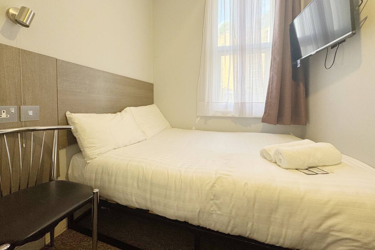 A hotel room with a bed, a desk, a TV mounted on the wall, and a window with curtains.