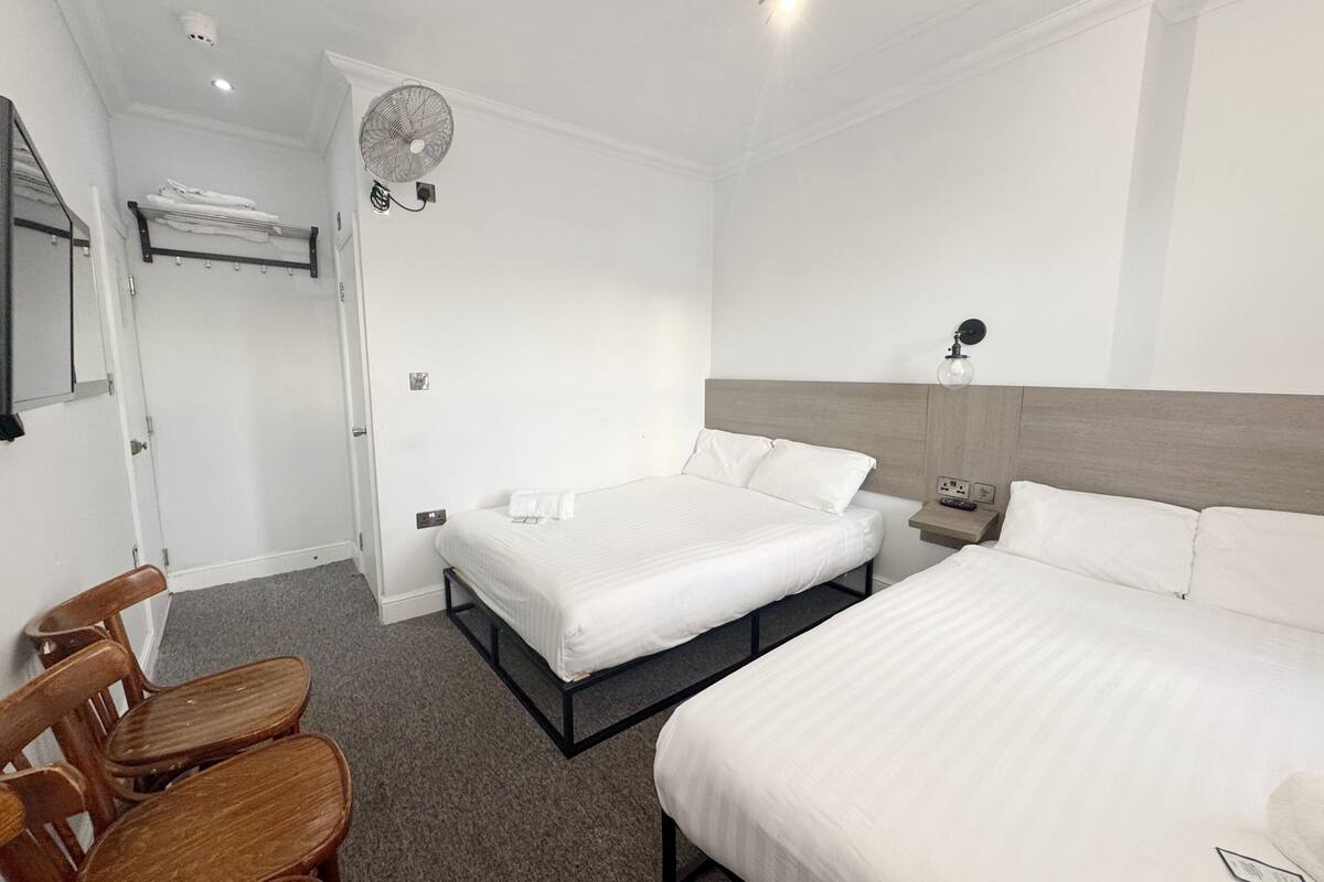 A hotel room with a large bed, a bedside table, a chair, a fan, and a wall-mounted light fixture.