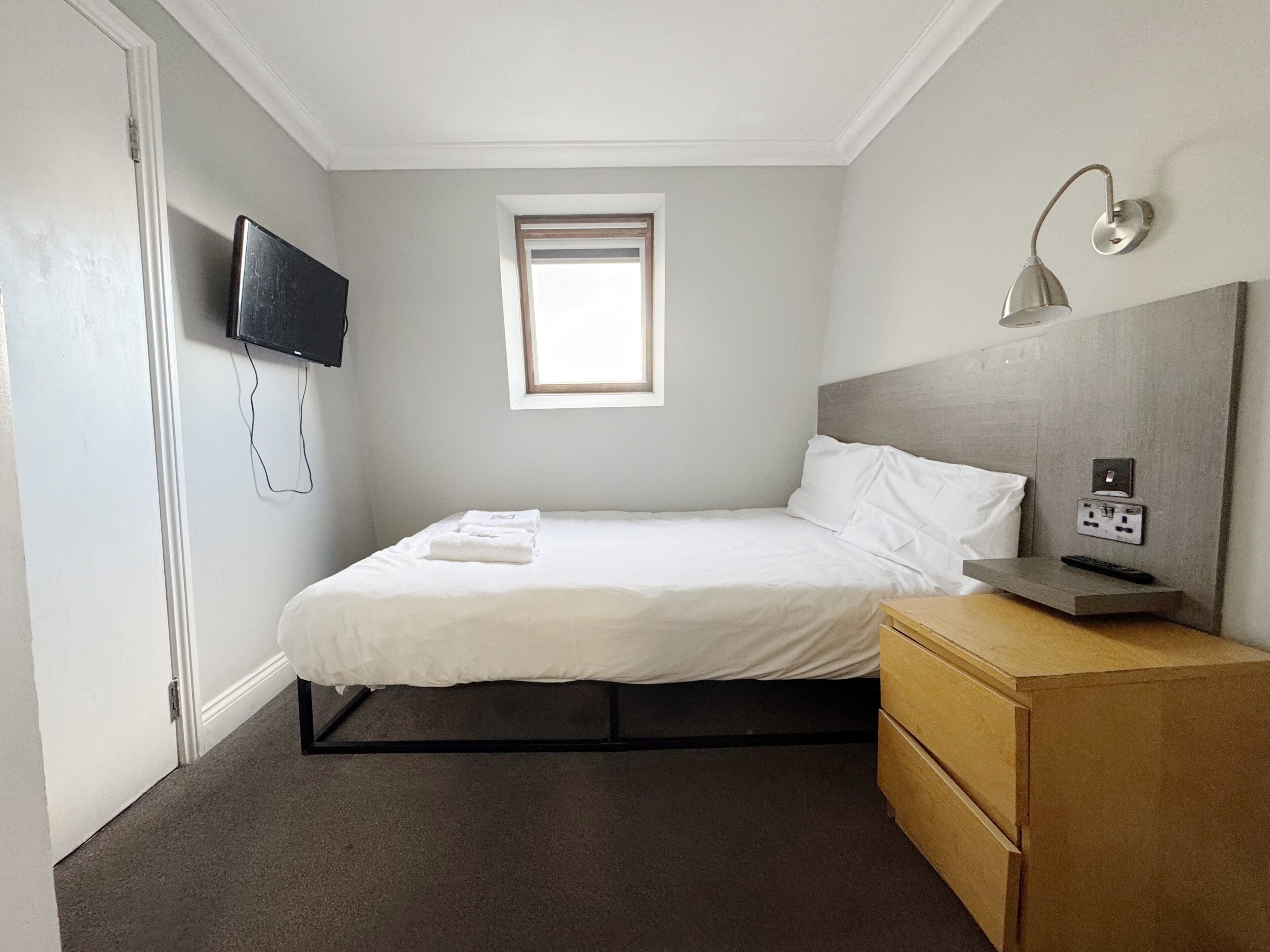A bedroom with a bed, a TV mounted on the wall, a small window, a bedside table with a lamp, and a power outlet.