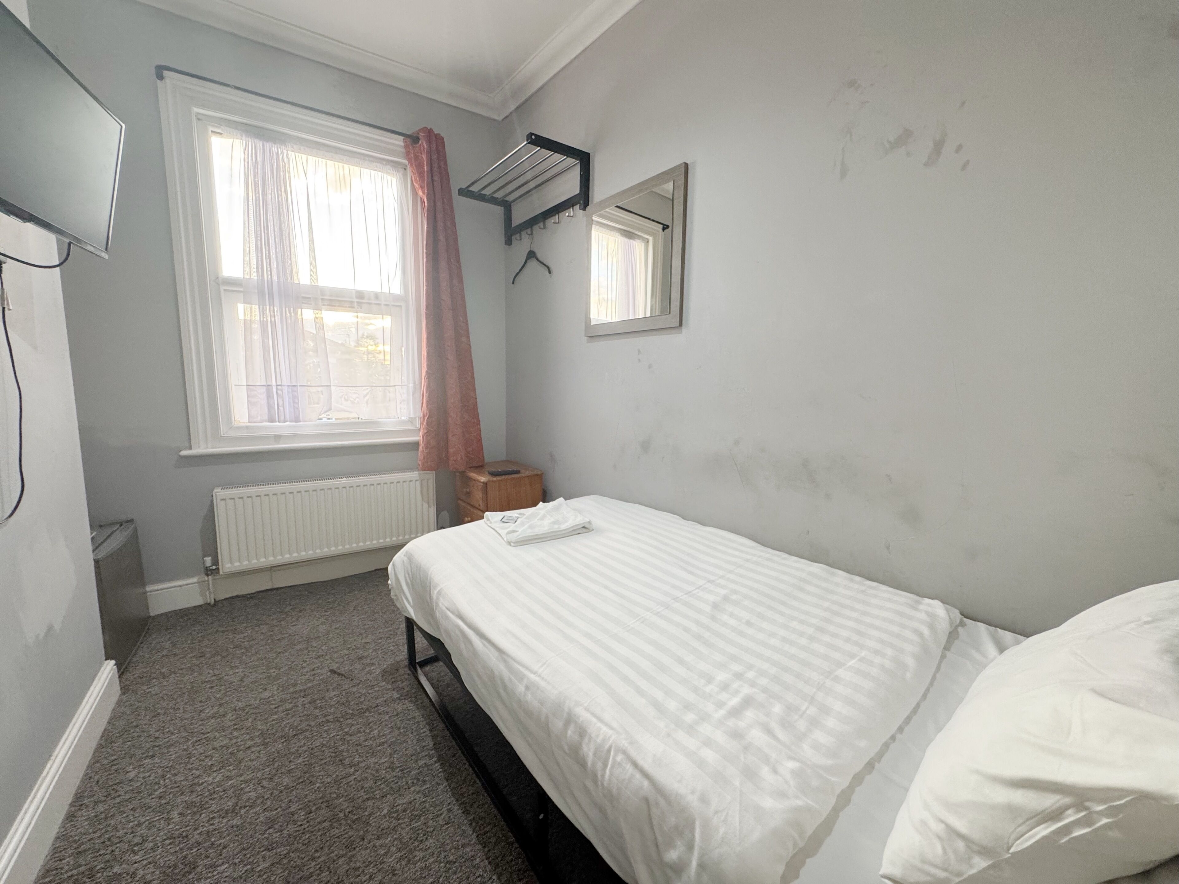 A bedroom with a single bed, a radiator, a window with curtains, a hanging clothes rack, and a mirror on the wall.