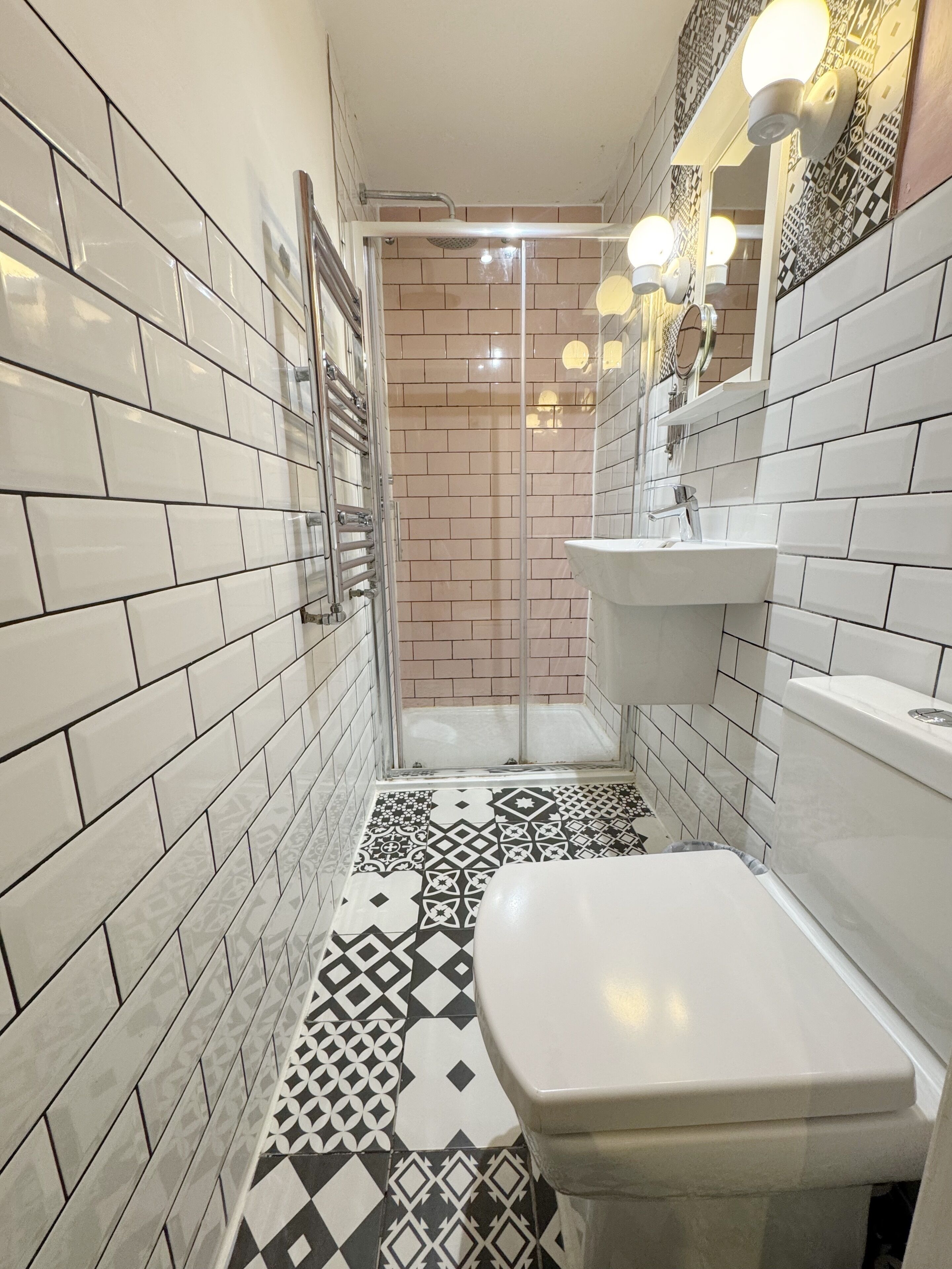 Bagno con piastrelle bianche, pavimento decorato, un water, un lavandino e una doccia con porta in vetro.