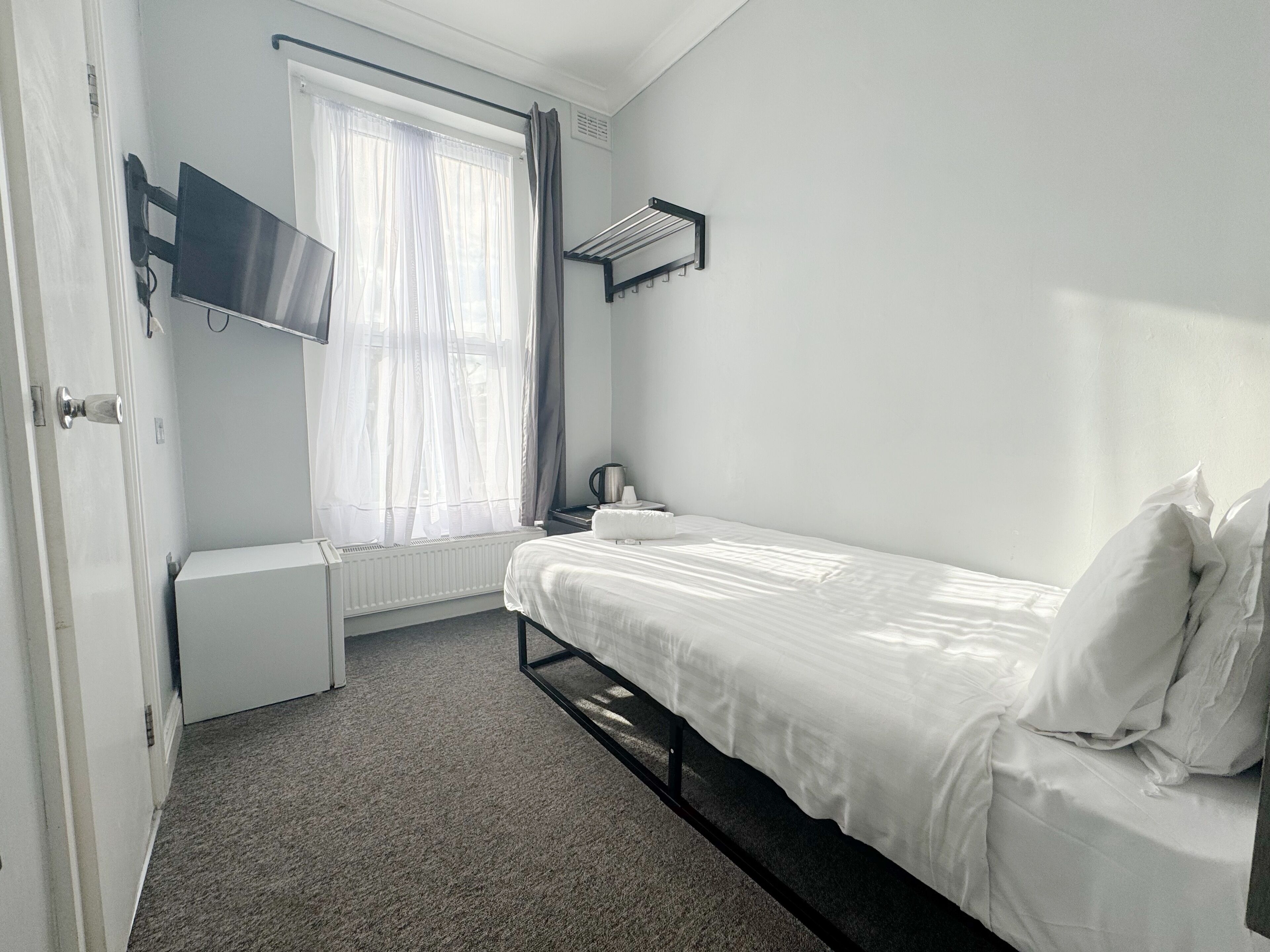 A small, clean room with a bed, a radiator, a TV mounted on the wall, and a window with curtains.
