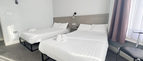 Ein Hotelzimmer mit zwei Betten, einem Standventilator, einem Schreibtisch und einem Fenster mit Vorhängen.