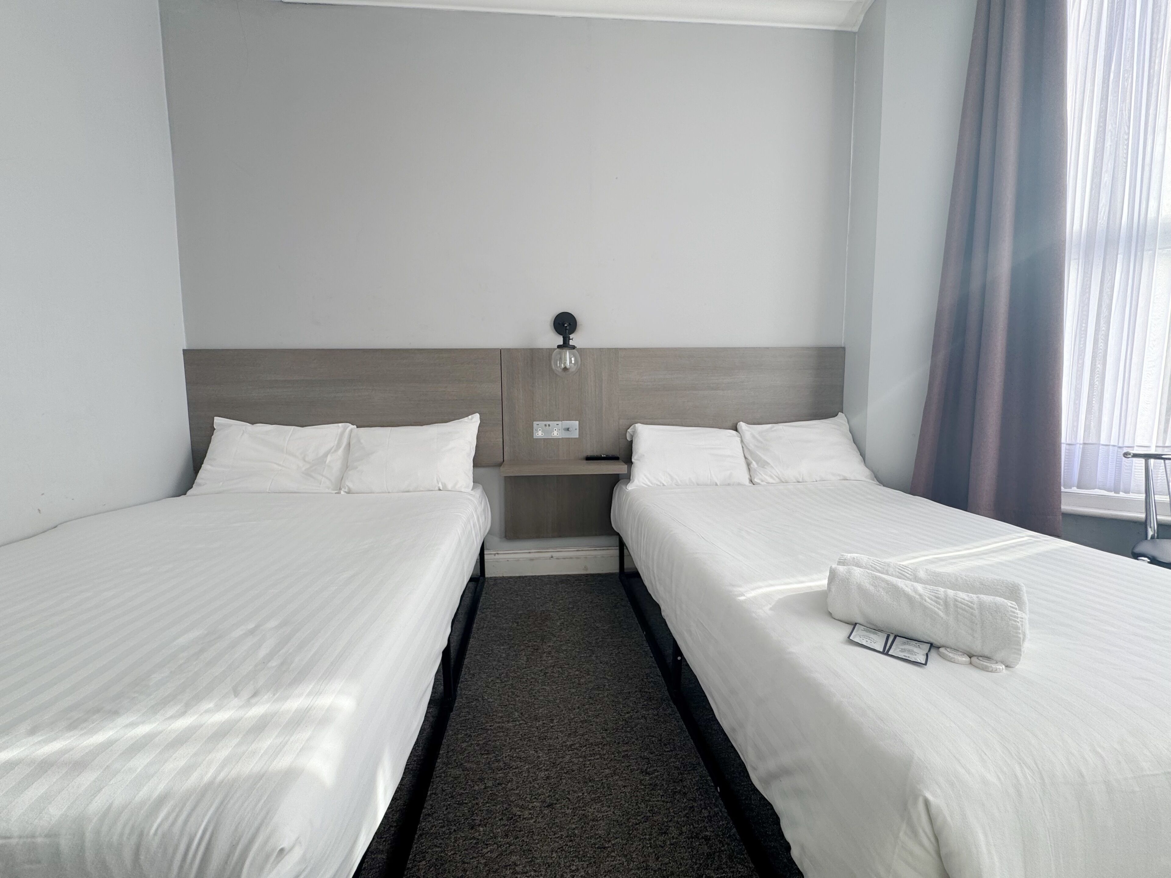 Une chambre d’hôtel avec deux lits, une table de chevet, une lampe fixée au mur et une fenêtre avec des rideaux.