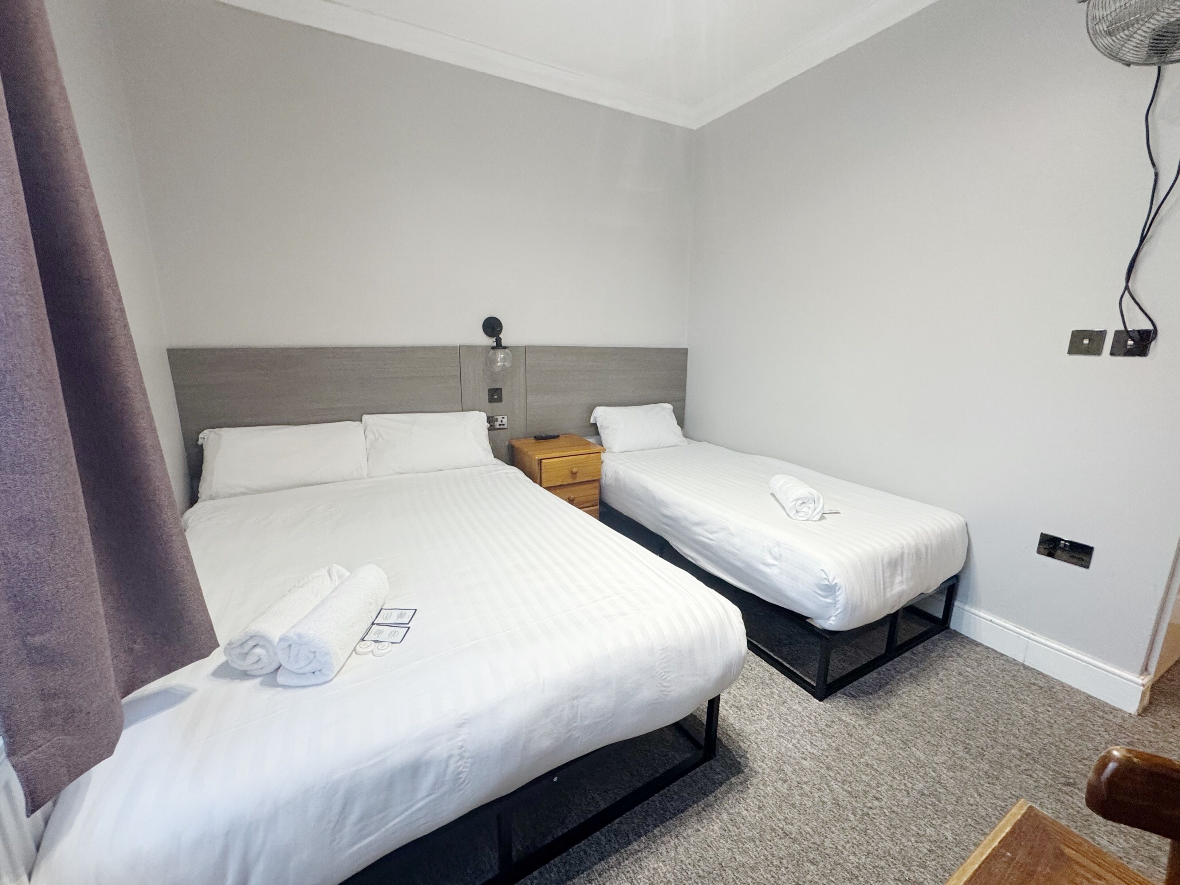 A hotel room with two single beds, a wooden nightstand, a wall-mounted fan, and a power outlet.