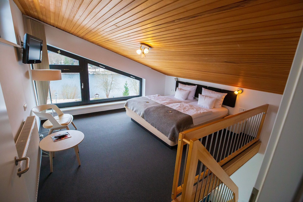 A modern bedroom with a large bed, a desk, and a view of the outdoors through a window.