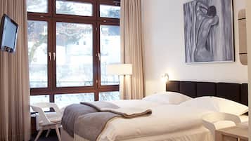 Quarto de hotel moderno com uma cama grande, uma escrivaninha e uma cadeira. Há uma janela com cortinas, um quadro na parede e uma televisão montada.
