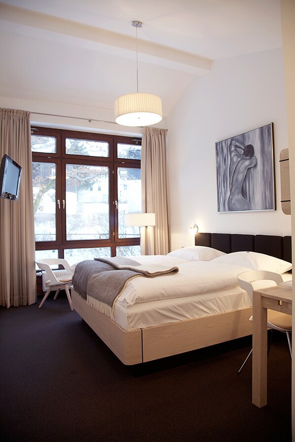 Una habitación de hotel moderna con una cama grande, un escritorio y una silla. Hay una ventana con cortinas, un cuadro en la pared y un televisor montado.