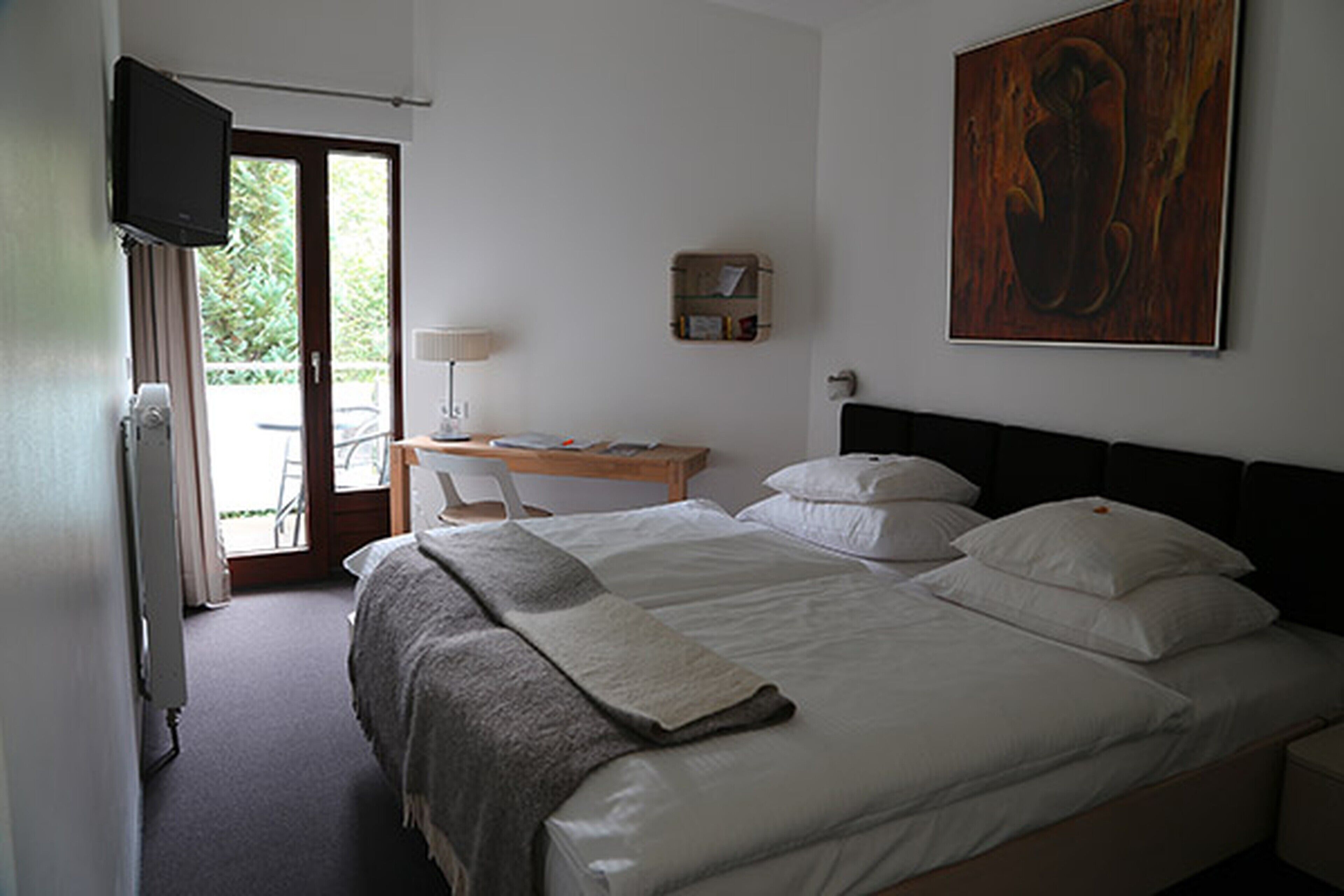A bedroom with a bed, a desk, a chair, a television, and a painting on the wall.