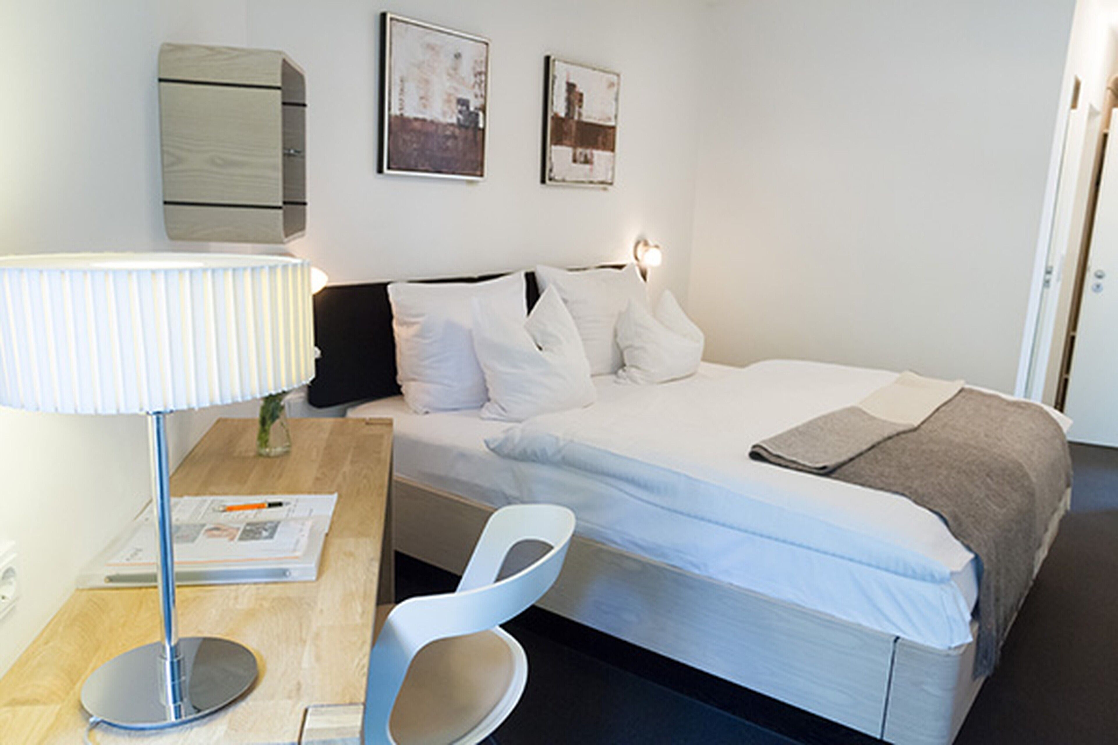Une chambre d’hôtel bien rangée, avec un lit, un bureau doté d’une lampe, une chaise et deux tableaux encadrés accrochés au mur.
