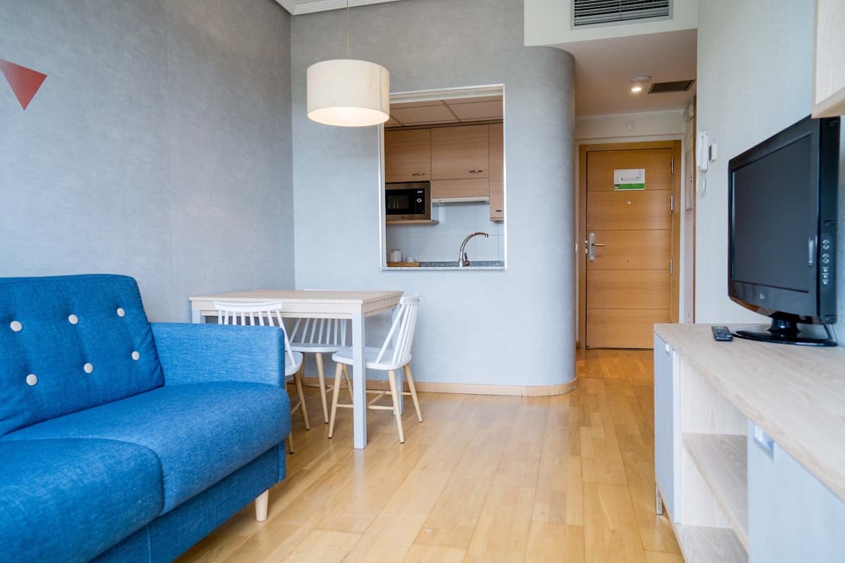 A modern living room with a blue sofa, a dining table with chairs, a kitchen area visible through an open doorway, and a television on a wooden stand.
