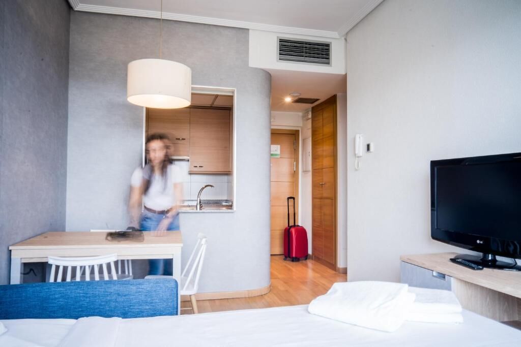 Habitación de hotel con cama, escritorio, televisor y kitchenette, además de una maleta roja.
