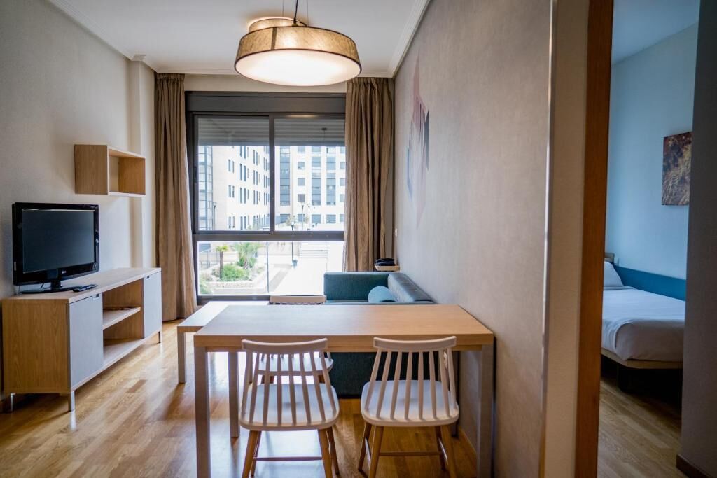 A modern hotel room with a wooden dining table, two white chairs, a television on a wooden stand, a large window with a view of buildings, and a bed with a blue headboard.