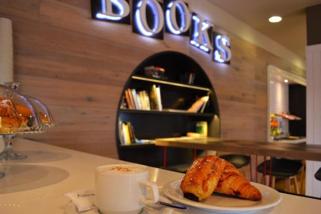 Una mesa con un croissant y una taza de café, una estantería y un cartel que dice 'BOOKS' al fondo.