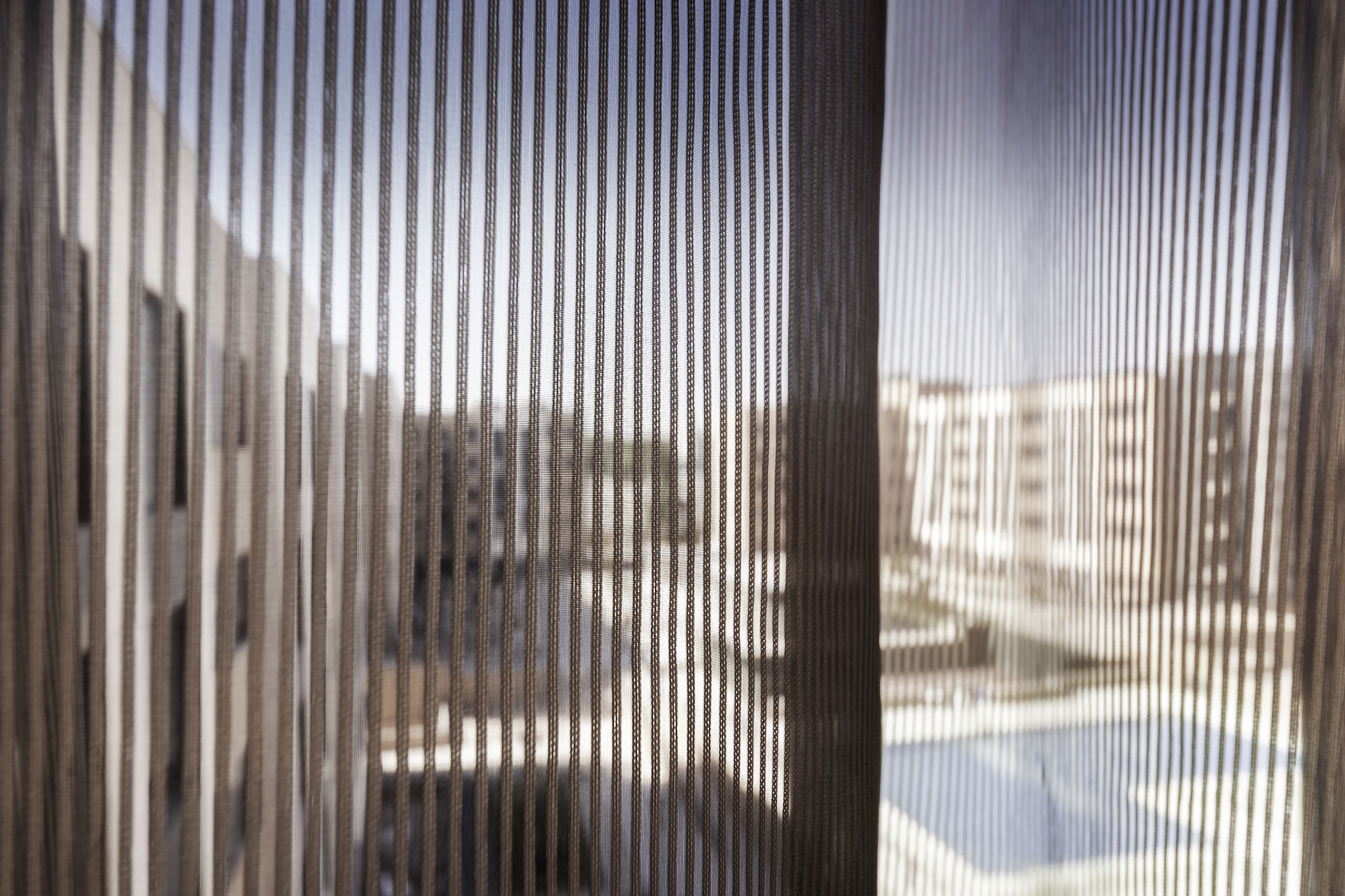Vista a través de una ventana con persianas verticales, mostrando un edificio de varios pisos y un cielo despejado.