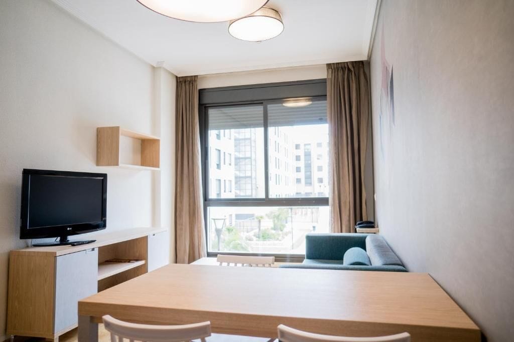 Ett modernt rum med en platt-TV, ett matbord i trä, två stolar, en soffa och ett stort fönster med gardiner.
