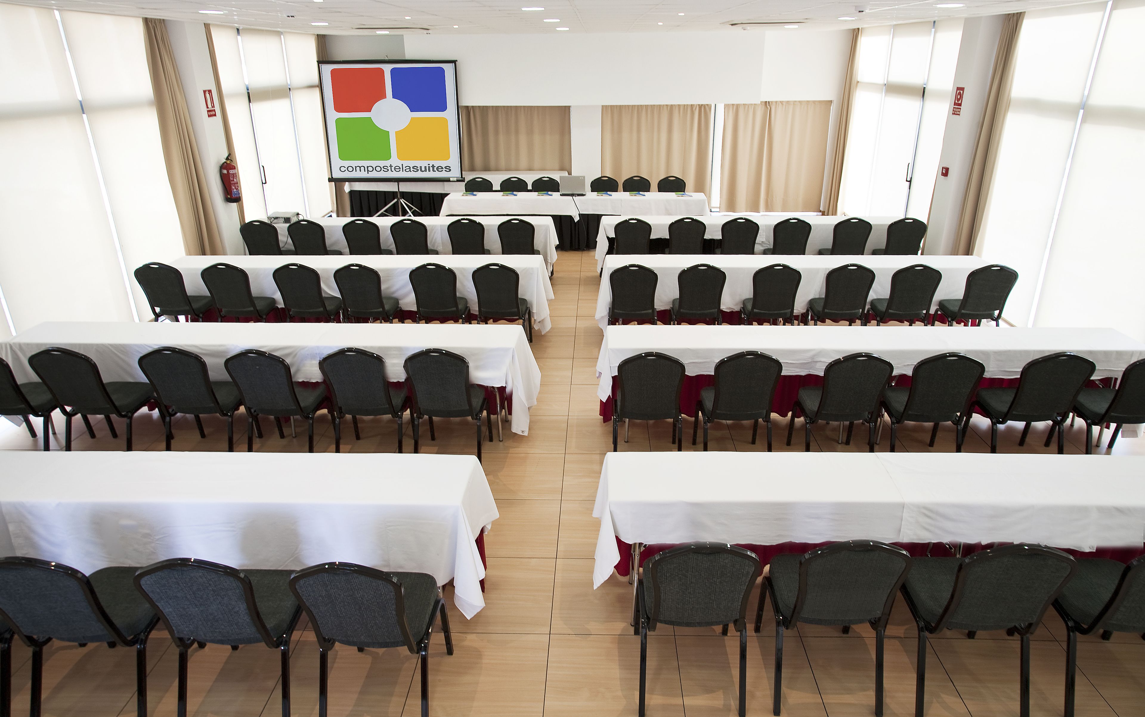 Une salle de conférence équipée de plusieurs tables et chaises, d’un écran de projection et d’un extincteur.