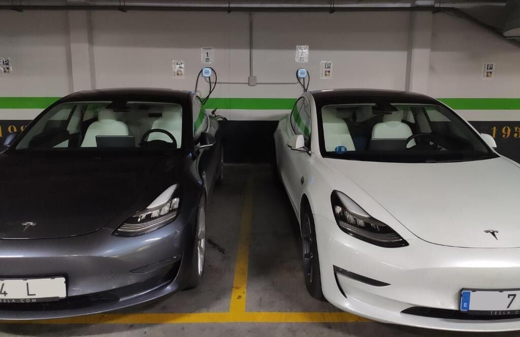 Two Tesla cars parked side by side in a parking garage with green and white walls, and a yellow line marking the parking spaces.