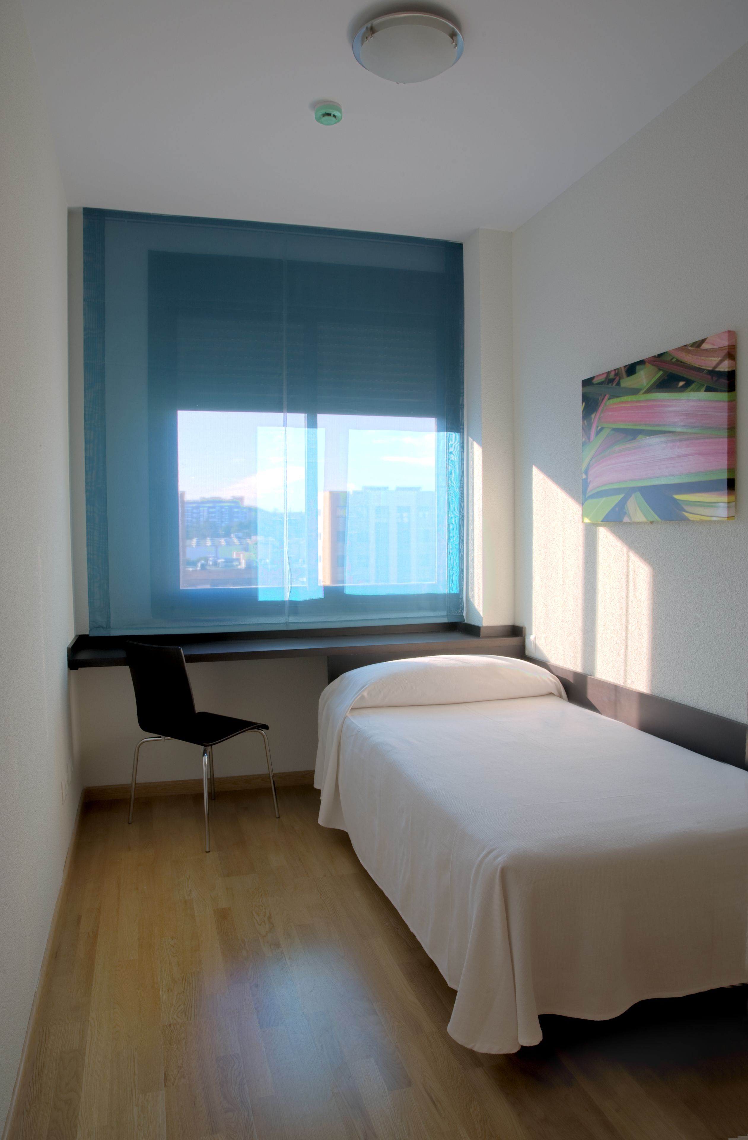 A hotel room with a large window, a bed, a desk, and a chair.