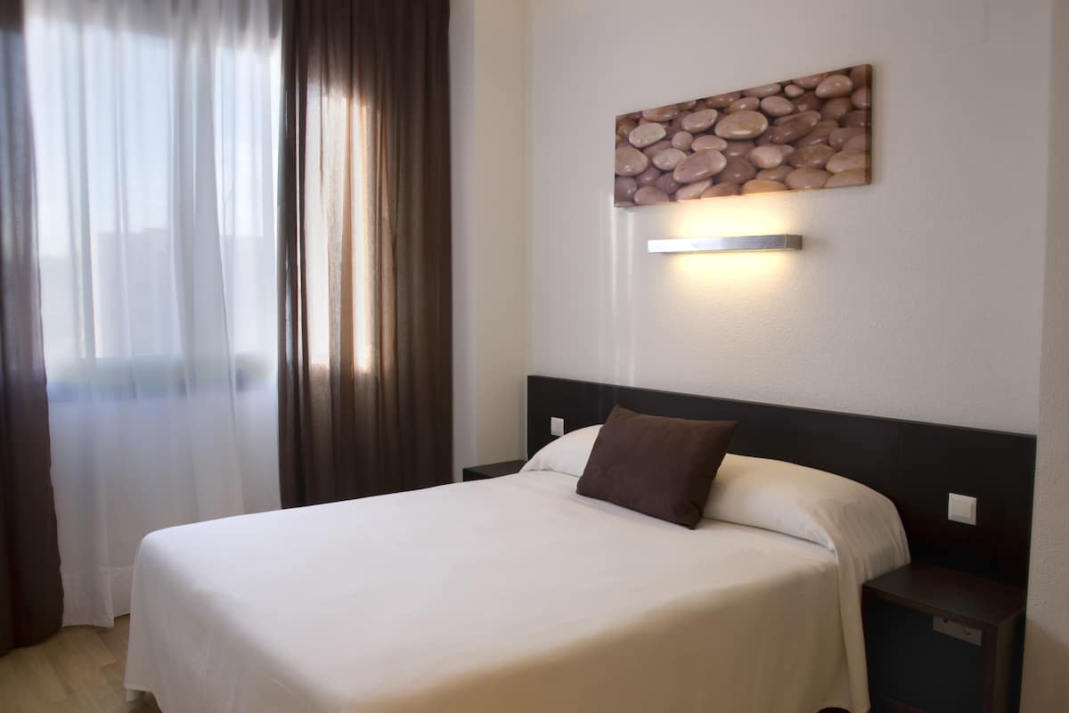 A hotel room with a large bed, a nightstand, a wall-mounted light, and a window with curtains.