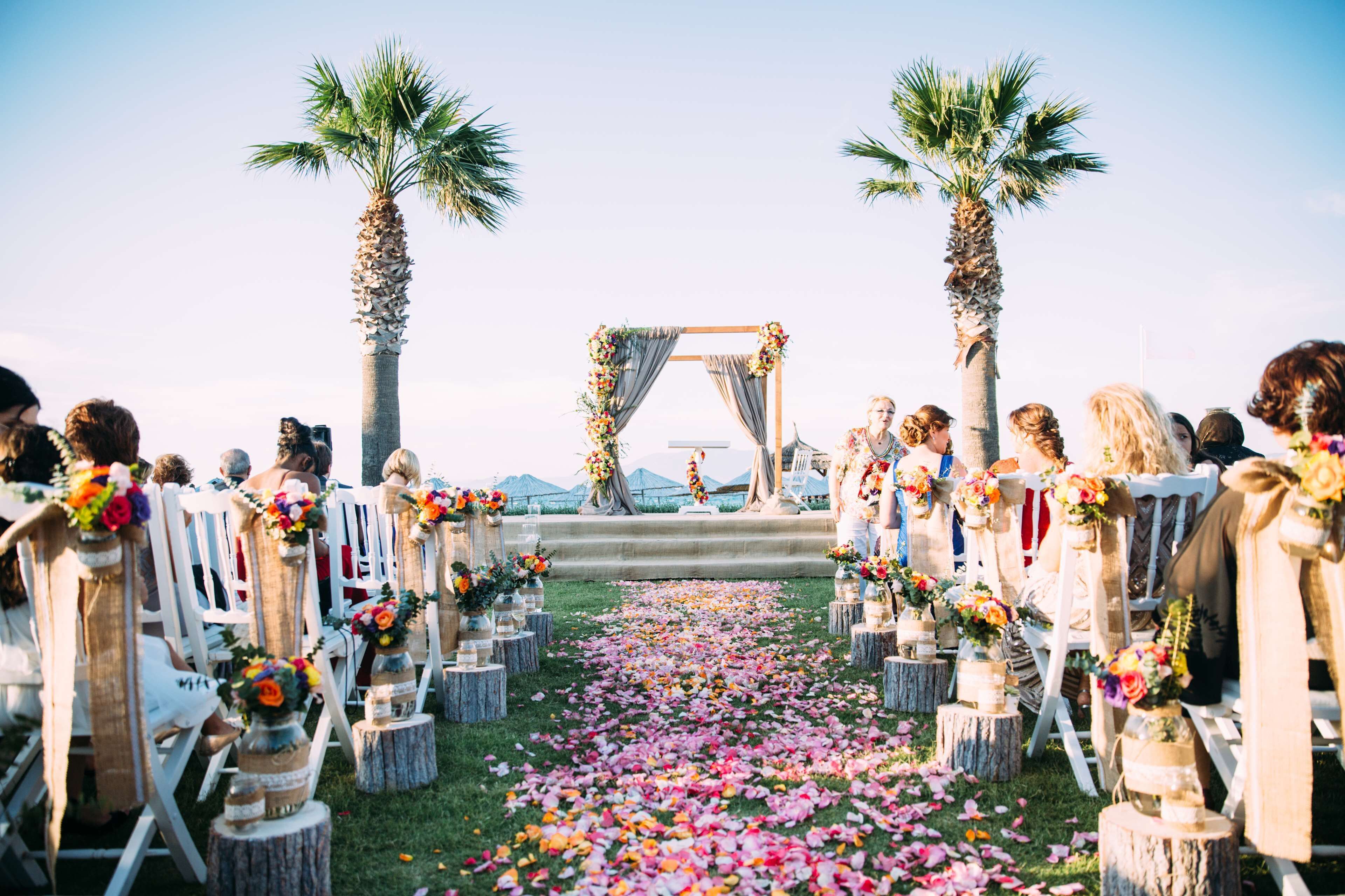 A wedding ceremony aisle with white chairs, floral decorations, and palm trees.