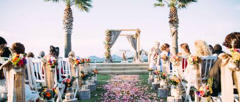 Pasillo de una ceremonia de boda con sillas blancas, adornos florales y palmeras.