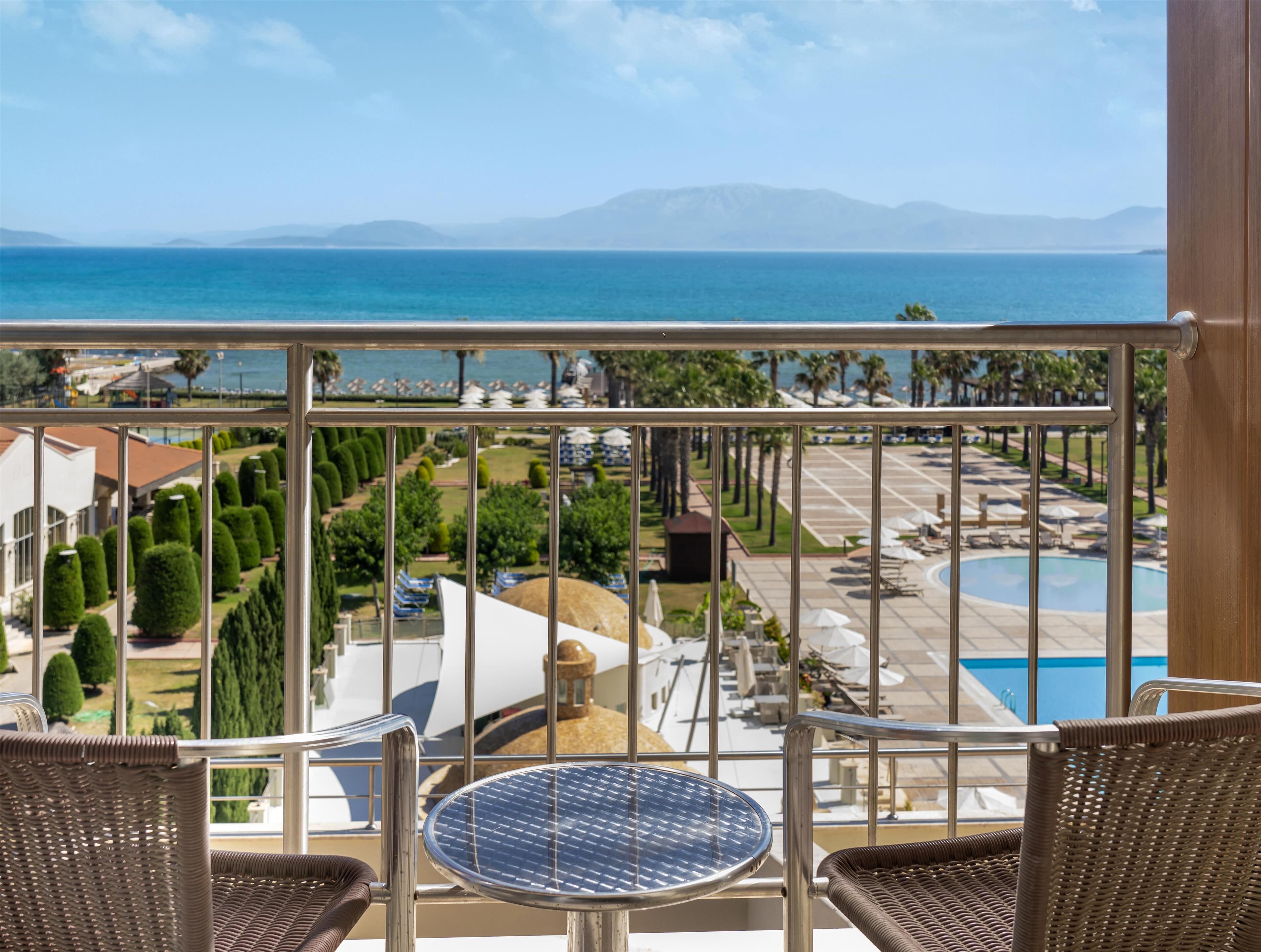 A balcony with wicker chairs and a round table, overlooking a pool and a landscaped area with palm trees and a view of the sea and mountains.