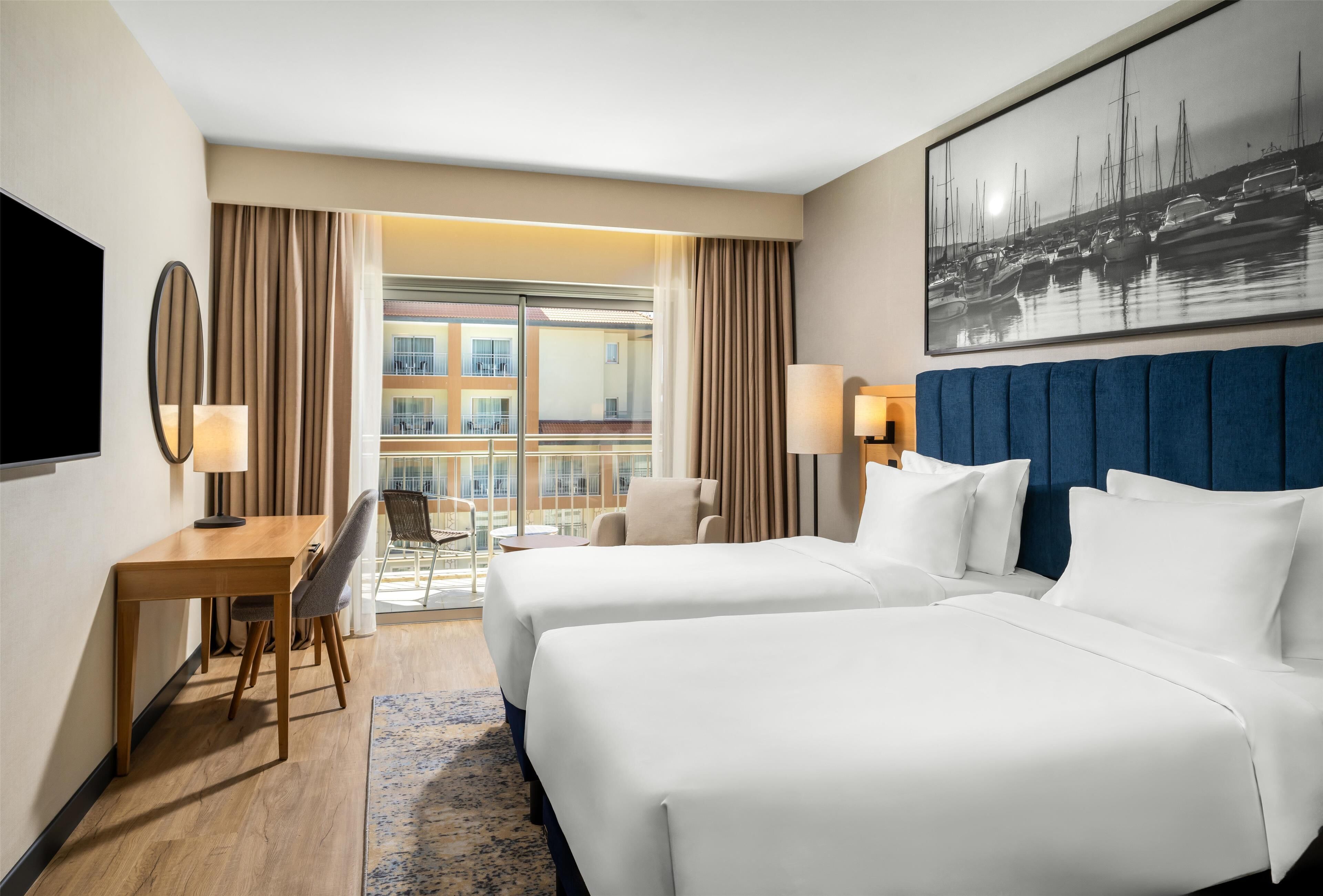 A hotel room with two beds, a desk with a chair, a TV, and a view of a building and boats through the window.