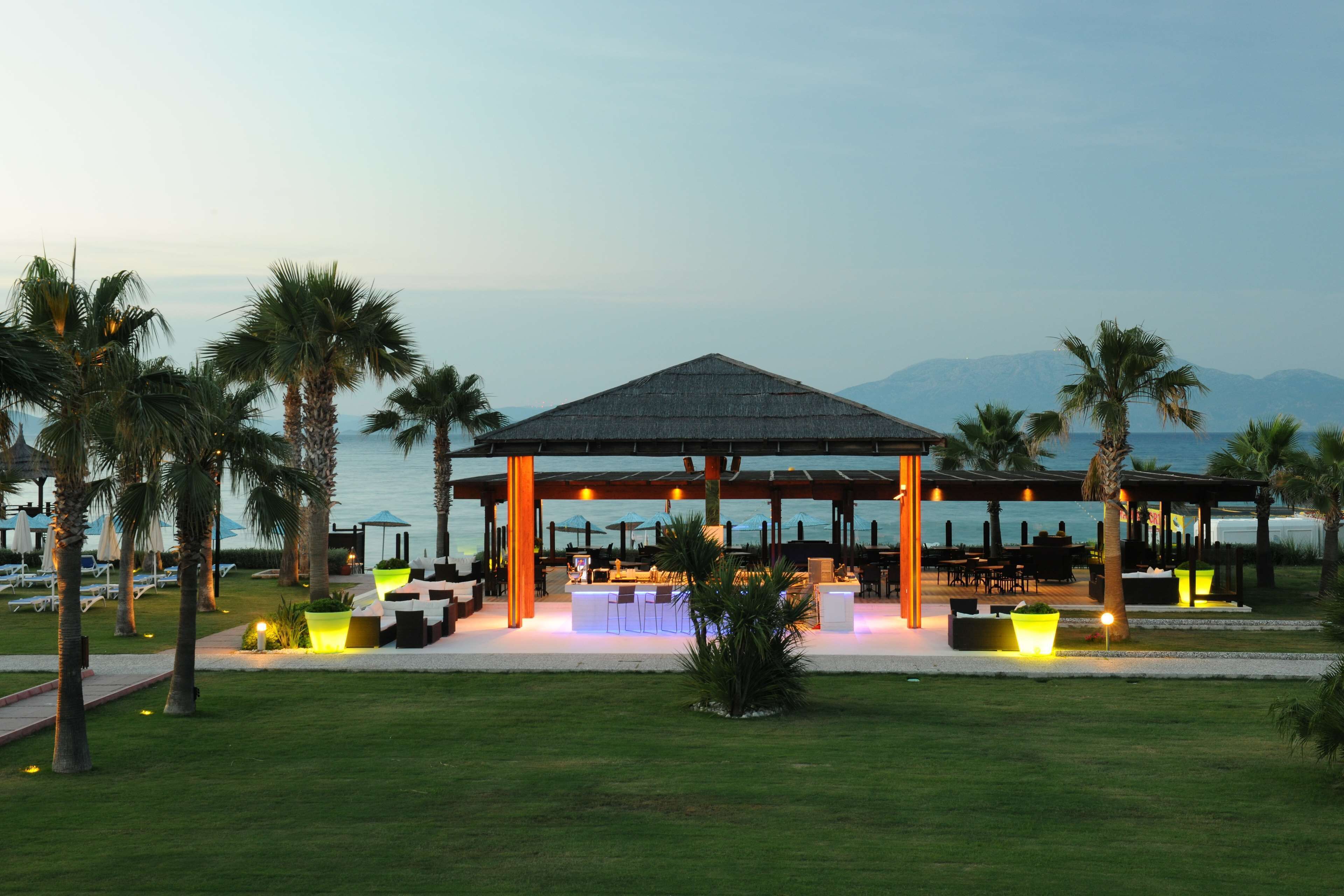 Un restaurante frente al mar con terraza, palmeras y vistas al océano y las montañas.