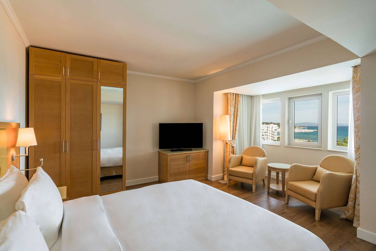 Ein Hotelzimmer mit Bett, Fernseher, Schreibtisch, zwei Stühlen und Meerblick.