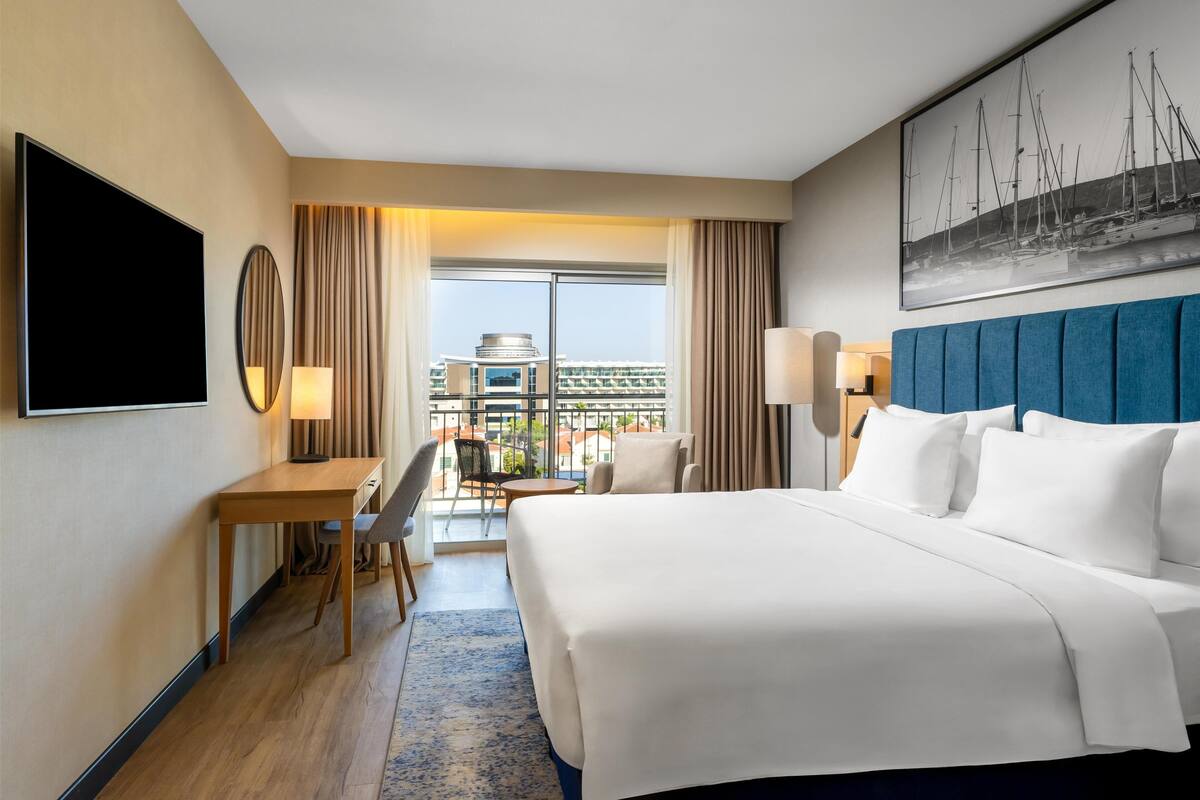 Ein Hotelzimmer mit einem großen Bett, einem Schreibtisch mit Stuhl, einem Fernseher und Blick auf Gebäude durch das Fenster.
