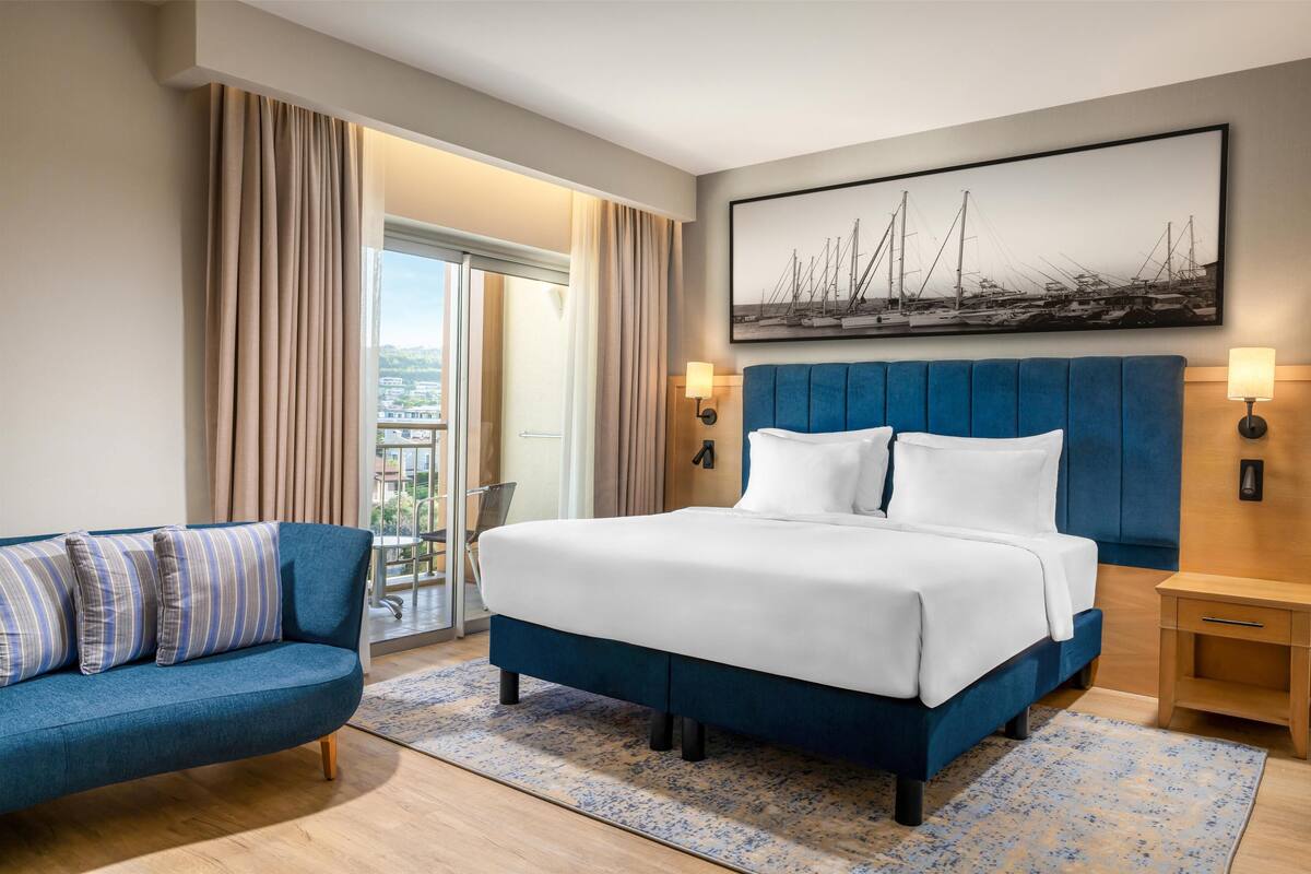 Ein Hotelzimmer mit Bett, einer Couch, Blick auf die Stadt und einem Gemälde an der Wand.