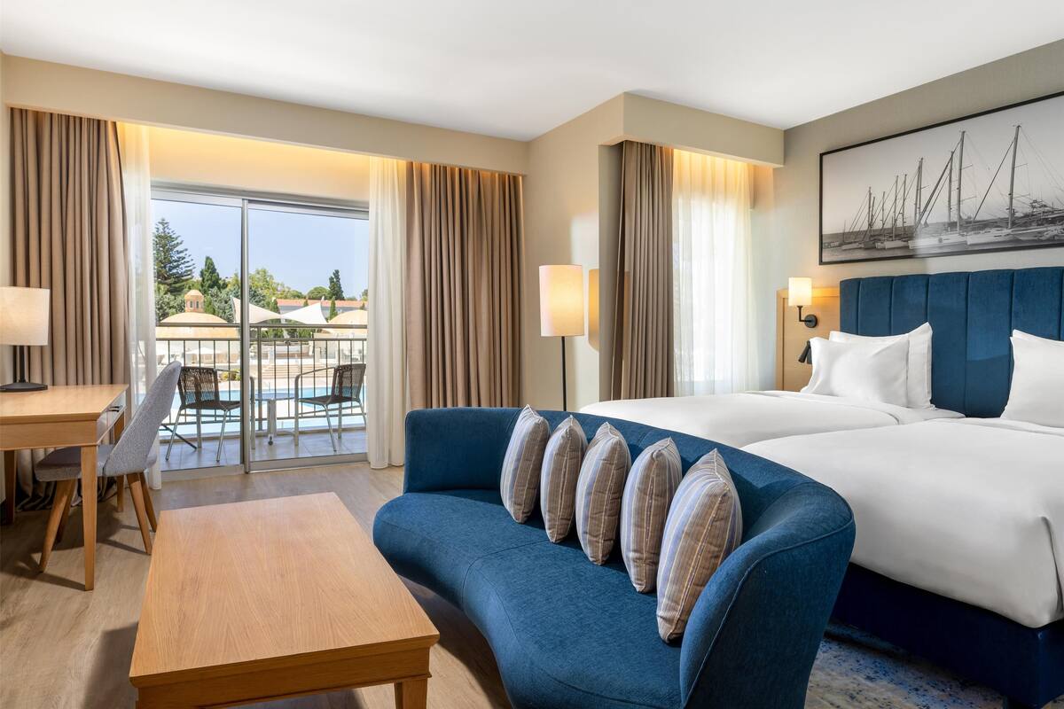 Ein Hotelzimmer mit einem Bett, einem blauen Sofa, einem Holztisch und Blick ins Freie durch eine Glastür.