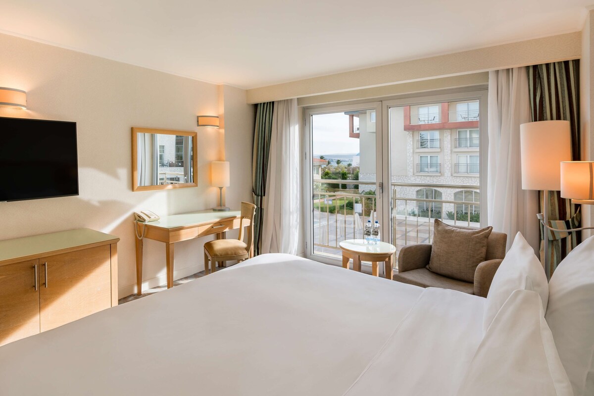 Hotellihuone, jossa on sänky, työpöytä, tuoli, pieni pöytä, televisio ja ikkuna, josta on näkymä rakennuksiin.