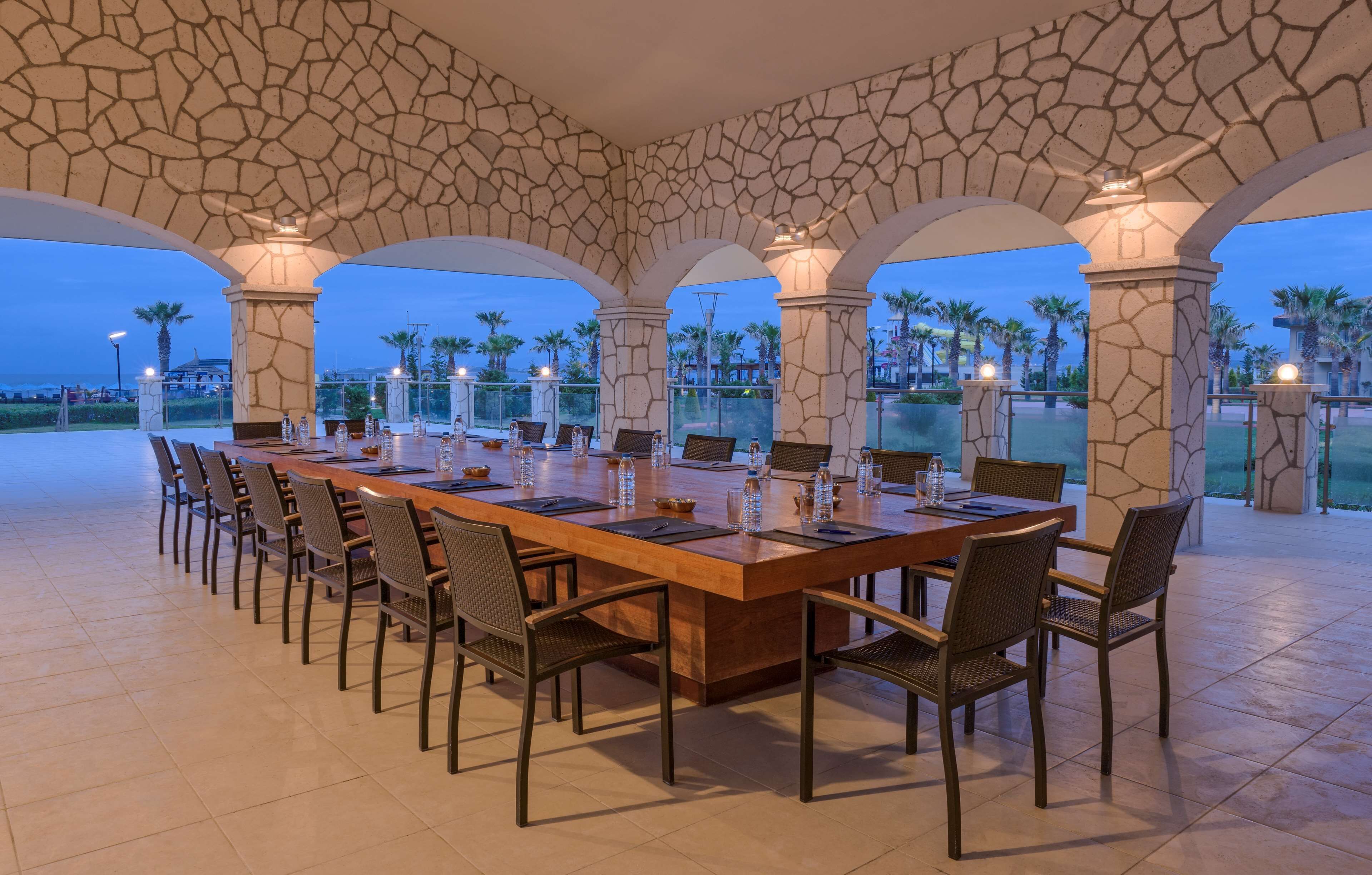 A long dining table with chairs, set in a spacious outdoor area with stone arches and palm trees in the background.
