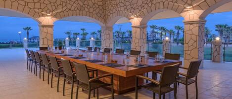 A long dining table with chairs, set in a spacious outdoor area with stone arches and palm trees in the background.