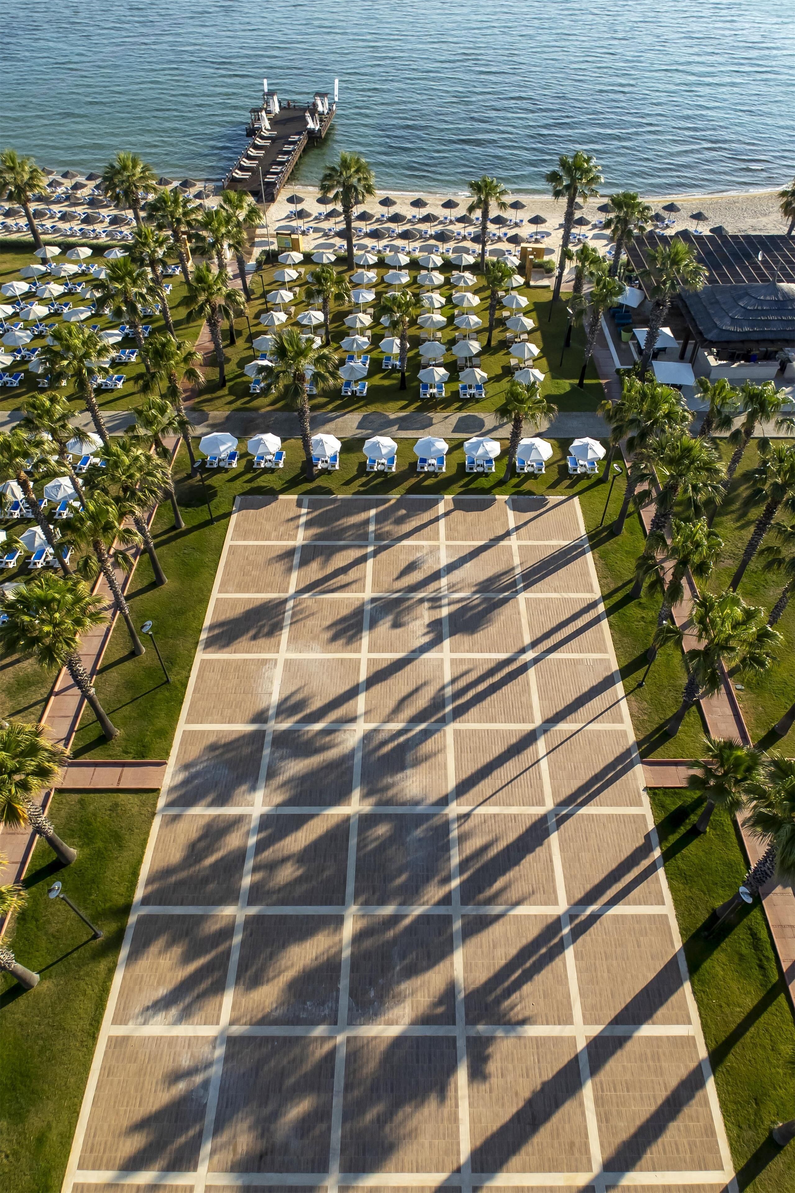 An aerial view of a beachside resort with a large open deck area, palm trees, and beach umbrellas.