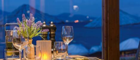 A dining table set with wine glasses, a candle, and a vase of flowers, overlooking a mountainous landscape.