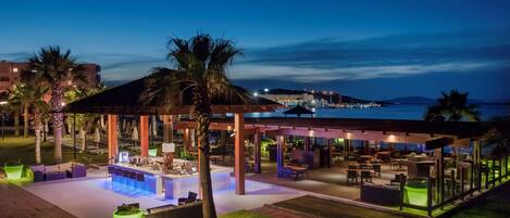 An outdoor lounge area with seating and a bar, surrounded by palm trees and a view of the sea.