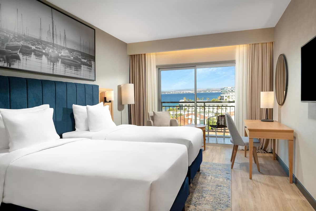 Ein Hotelzimmer mit zwei Betten, einem Schreibtisch, einem Stuhl, einem Fernseher und einem großen Fenster mit Blick auf das Meer und Gebäude.