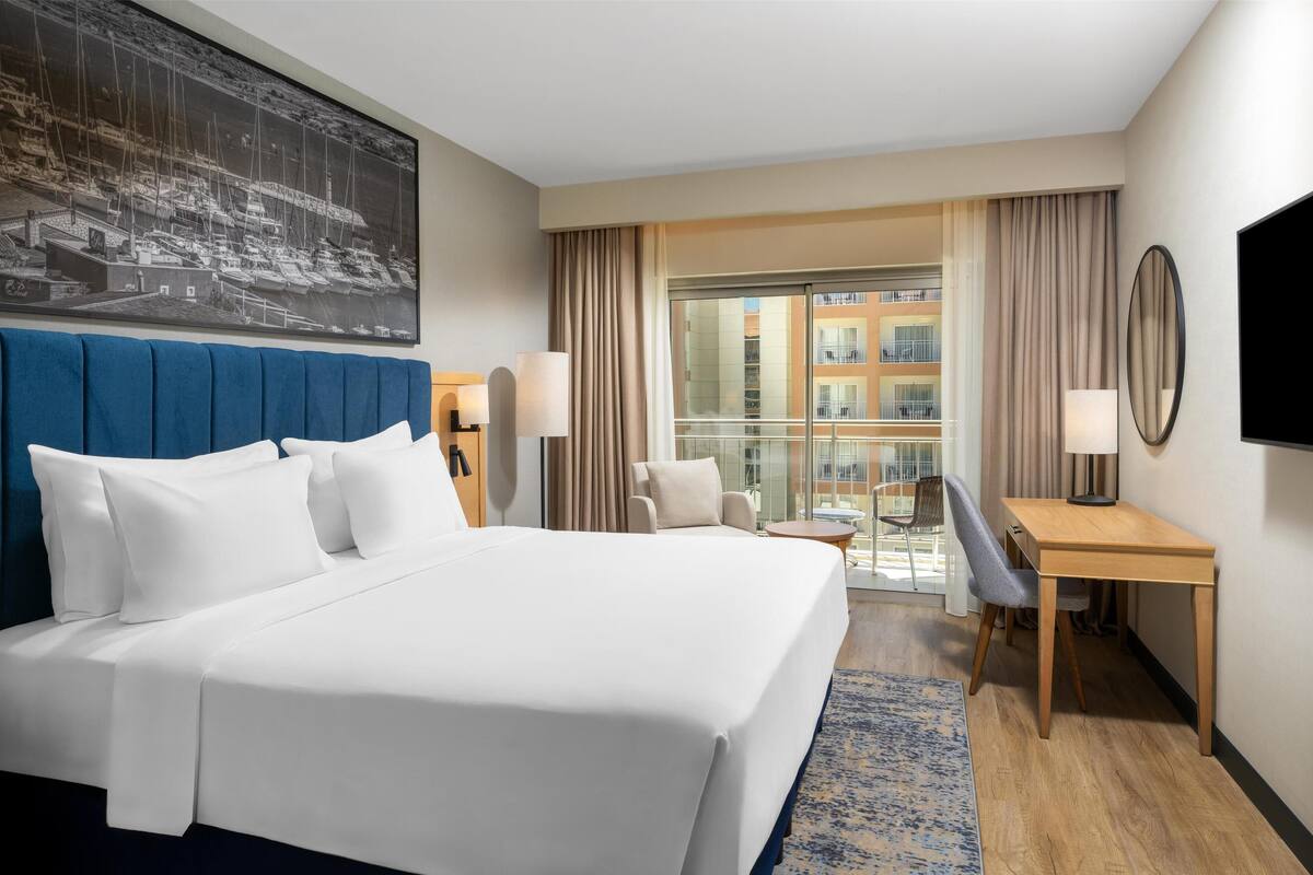 Ein Hotelzimmer mit einem großen Bett, einem Schreibtisch, einem Stuhl, einem Fernseher und einem Balkon mit Blick auf Gebäude.